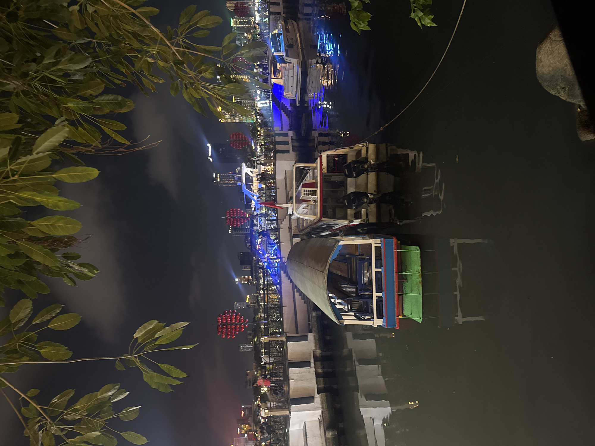 This is a night scene in Đà Nẵng, Vietnam. The image is dominated by the river, with several boats docked along the pier. One particular boat features a blue and red trim with a green tarp-like structure covering the deck. In the background, the cityscape is brightly lit with various buildings and structures, including what appear to be red lantern decorations. On the left side of the frame, tree foliage is visible. The reflection of the city lights and boats are captured in the water, creating a vibrant scene. The photo is taken from a slightly elevated viewpoint, looking down towards the river and the city.