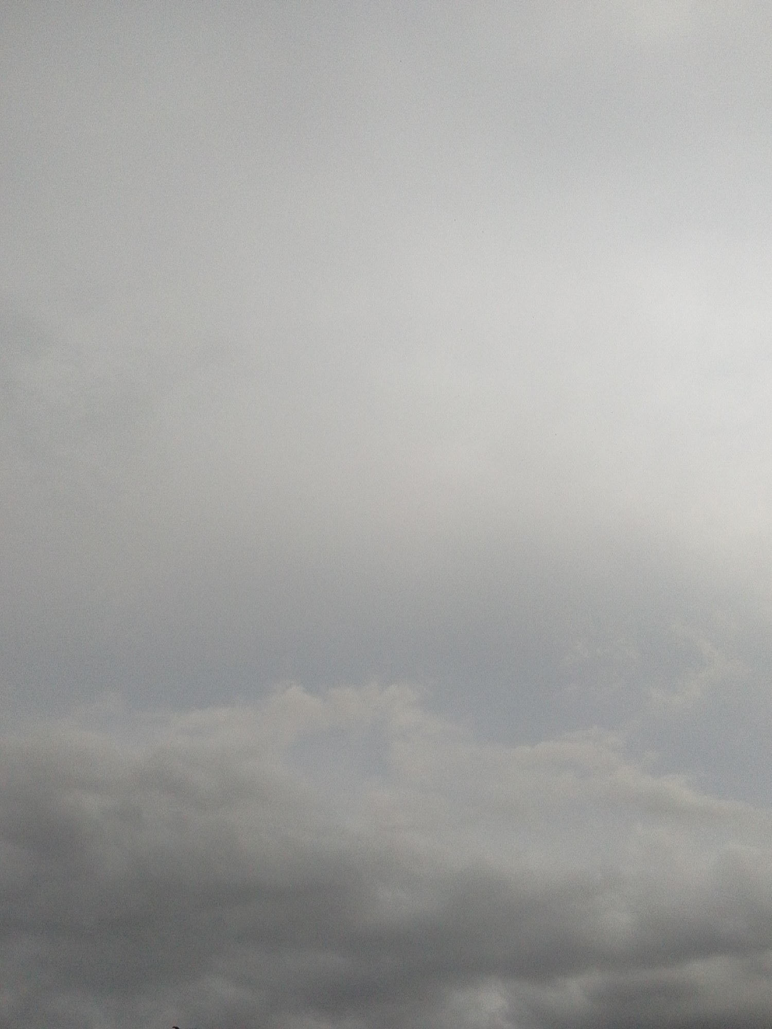 A high-angle medium shot captures a sky filled with overcast clouds. The clouds are a mix of light and dark gray, with some areas appearing almost white due to the diffused light. The bottom portion of the image shows the darker, denser clouds, while the upper portion transitions to lighter, thinner clouds that blend into the bright sky. There are no discernible buildings or other structures visible in this image, and no people or objects are present. The scene is devoid of any specific environmental context beyond the sky and clouds. This image was taken in Serti, Nigeria.