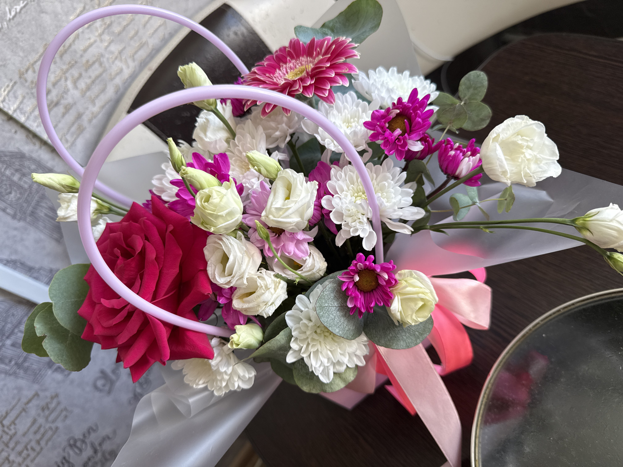 This is a close-up, top-down shot of a vibrant floral bouquet, possibly prepared for sale or as a gift. The bouquet features a prominent deep pink rose at the top, surrounded by white chrysanthemums, pink and white spray roses, and fuchsia-colored gerbera daisies. Accompanying the flowers are several sprigs of eucalyptus greenery. The arrangement is wrapped in translucent white paper and tied with light pink and hot pink ribbons. A decorative, light purple heart-shaped accessory is placed through the bouquet. The bouquet rests on a dark wooden surface. In the bottom left corner, a reflective, circular object, possibly a mirror or a metallic platter, is partially visible. The background consists of a grey textured surface with a subtle pattern of writing. There are no people or actions visible in the image. The scene is indoors, with the lighting suggesting it could be natural or artificial light. The location of the photograph is not specified, but the prompt indicates it is in the city of Vylygort, Russia.