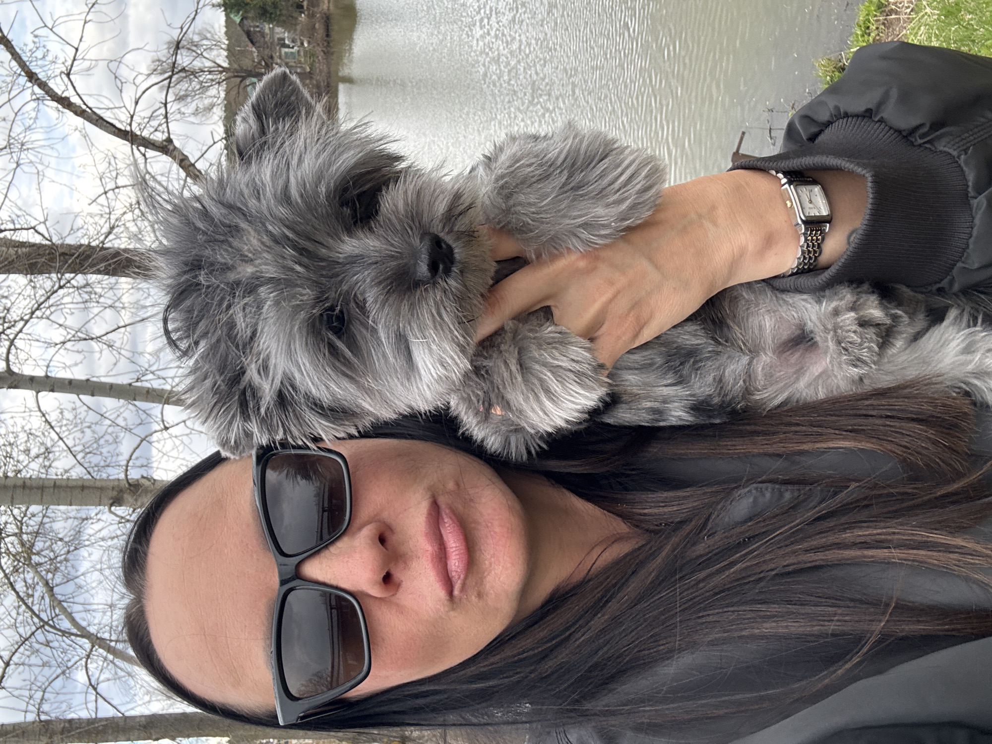 A person is holding a small, gray, shaggy-haired dog in an outdoor setting, likely captured in a selfie or close-up portrait. The image is oriented vertically, with the person's face and the dog's head prominent in the foreground.
The person has long, dark brown hair and is wearing black sunglasses, which obscure their eyes. Their lips are slightly parted, revealing a neutral expression. They are wearing a black jacket with ribbed cuffs. A silver-toned wristwatch with a rectangular face and a metal link band is visible on their left wrist.
The dog has dense, gray fur and small, dark eyes. It is looking directly towards the camera. The person's hand is visible supporting the dog.
In the background, there is a body of water, possibly a river or lake, with ripples on its surface. Beyond the water, there are bare trees, suggesting a season like late autumn, winter, or early spring. The sky overhead appears overcast.
No discernible text is legible on the wristwatch due to its size and resolution.