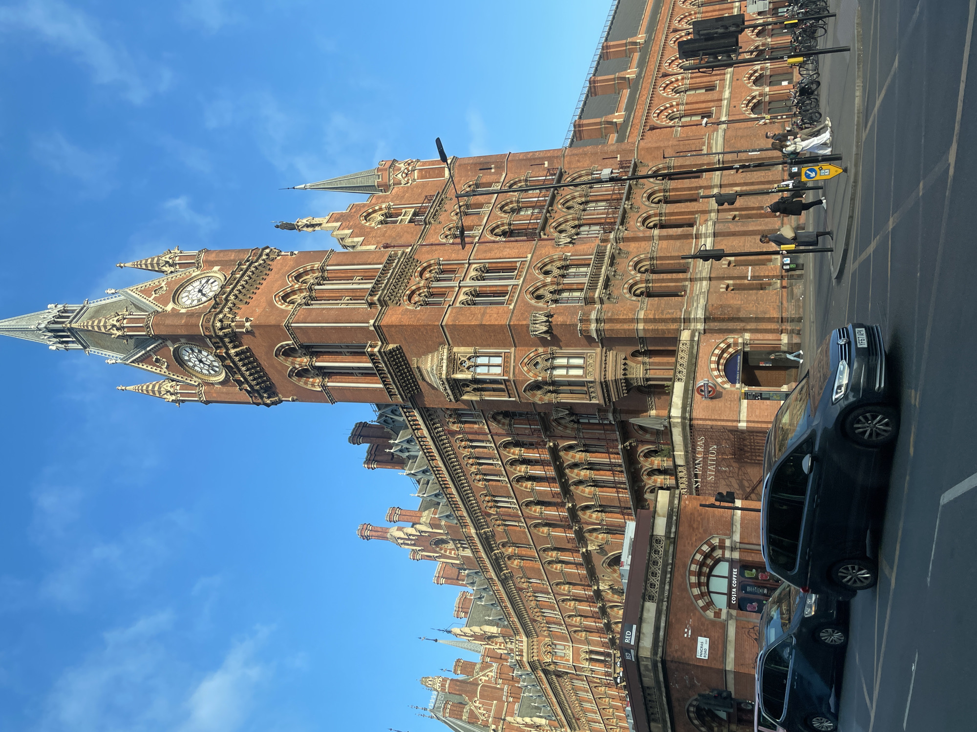 The image captures a view of St. Pancras Station in London, United Kingdom. The red-brick Victorian Gothic architecture of the station is prominently displayed, including its clock tower. Several parked cars line the street, partially obscuring the base of the building. Pedestrians can be seen walking along the sidewalk adjacent to the station. A clear blue sky is visible above the building. Street signs and lamp posts are also present along the street.