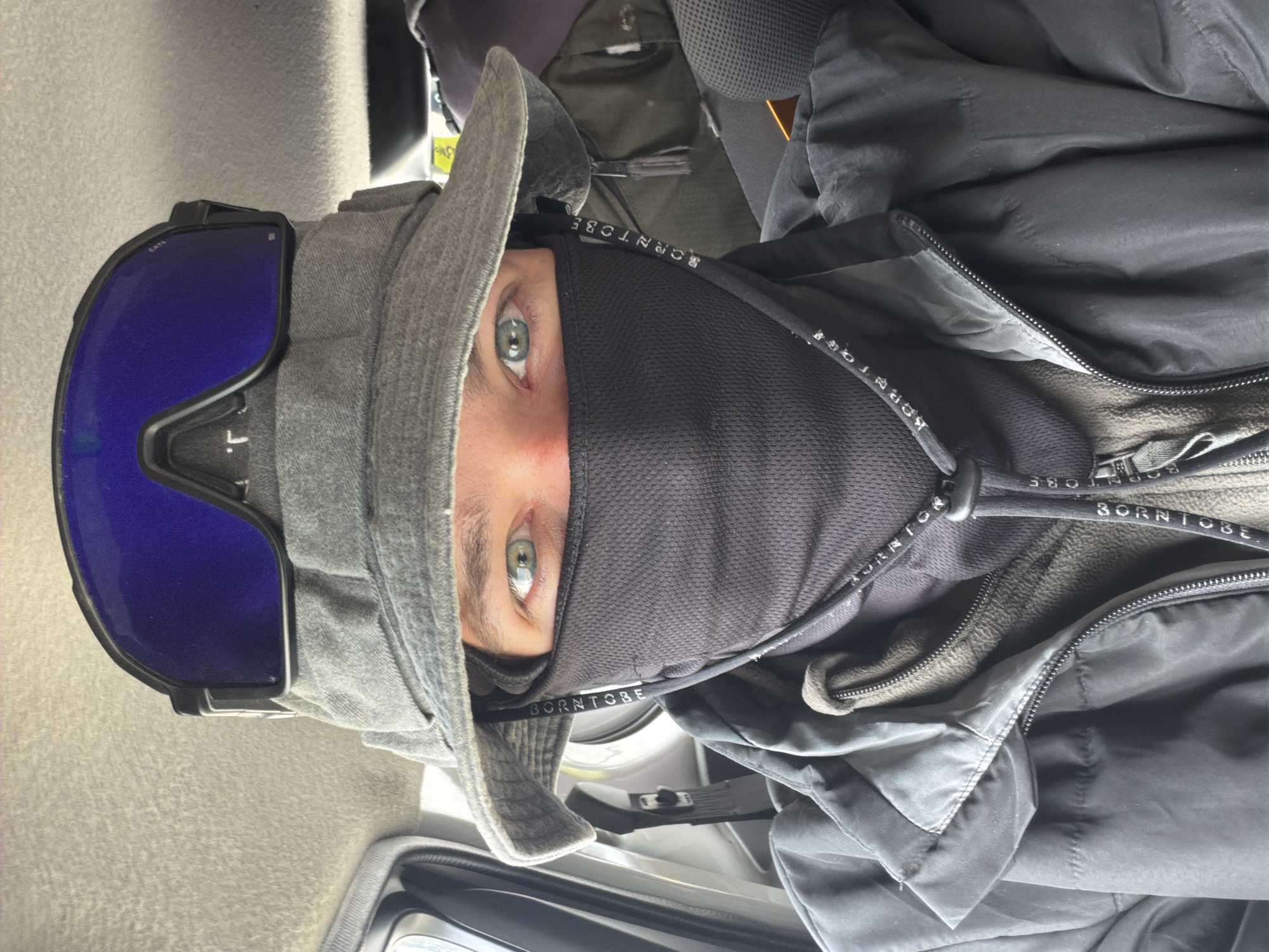 A person wearing a hat, goggles, and a face mask is inside a vehicle. The person has green eyes and is looking directly at the camera. The vehicle's interior is visible, with seats and a ceiling lining. The person is wearing a dark jacket over a fleece layer and a black neck gaiter. The goggles are blue and rest on top of the hat. The hat is a wide-brimmed bucket hat. The image does not provide enough information to determine the location within the city of Karzok, India.