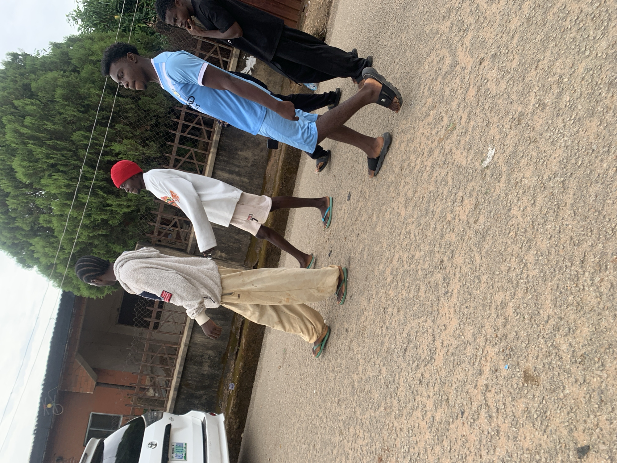 Three young men are walking on a paved road. The man closest to the viewer wears a light-colored hooded sweatshirt, beige trousers, and sandals. The second man wears a white sweater, light-colored shorts, and a red hat. The third man wears a blue soccer jersey with matching shorts and sandals. The background contains trees, a house, a fence, and a car. The image is taken in Jos, Nigeria.