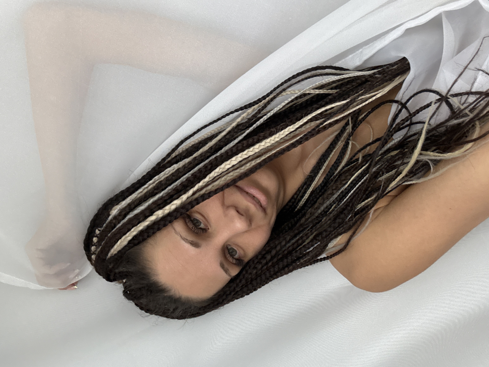 An artistic portrait captures a woman with long, dark braided hair, accentuated by interspersed light blonde or beige strands. She is depicted lying down, positioned diagonally across the frame, looking directly at the camera with a calm and serene expression, her lips slightly parted in a subtle smile. Her eyes are open, revealing their brown-hazel color and long dark eyelashes.
She is partially draped in, or lying on, soft, flowing translucent white fabric that covers much of the background and parts of her body. Her right arm, bent at the elbow, rests against this white surface, while her left leg and foot are faintly visible through the sheer material on the left side of the image. A glimpse of a light-colored toenail is visible on her left foot. The setting appears minimalist, likely indoors, with soft, even lighting illuminating her and the white fabric. No discernible text is present in the image. The scene suggests a relaxed photoshoot or intimate moment. The photo was taken in Rusanovo, Russia.