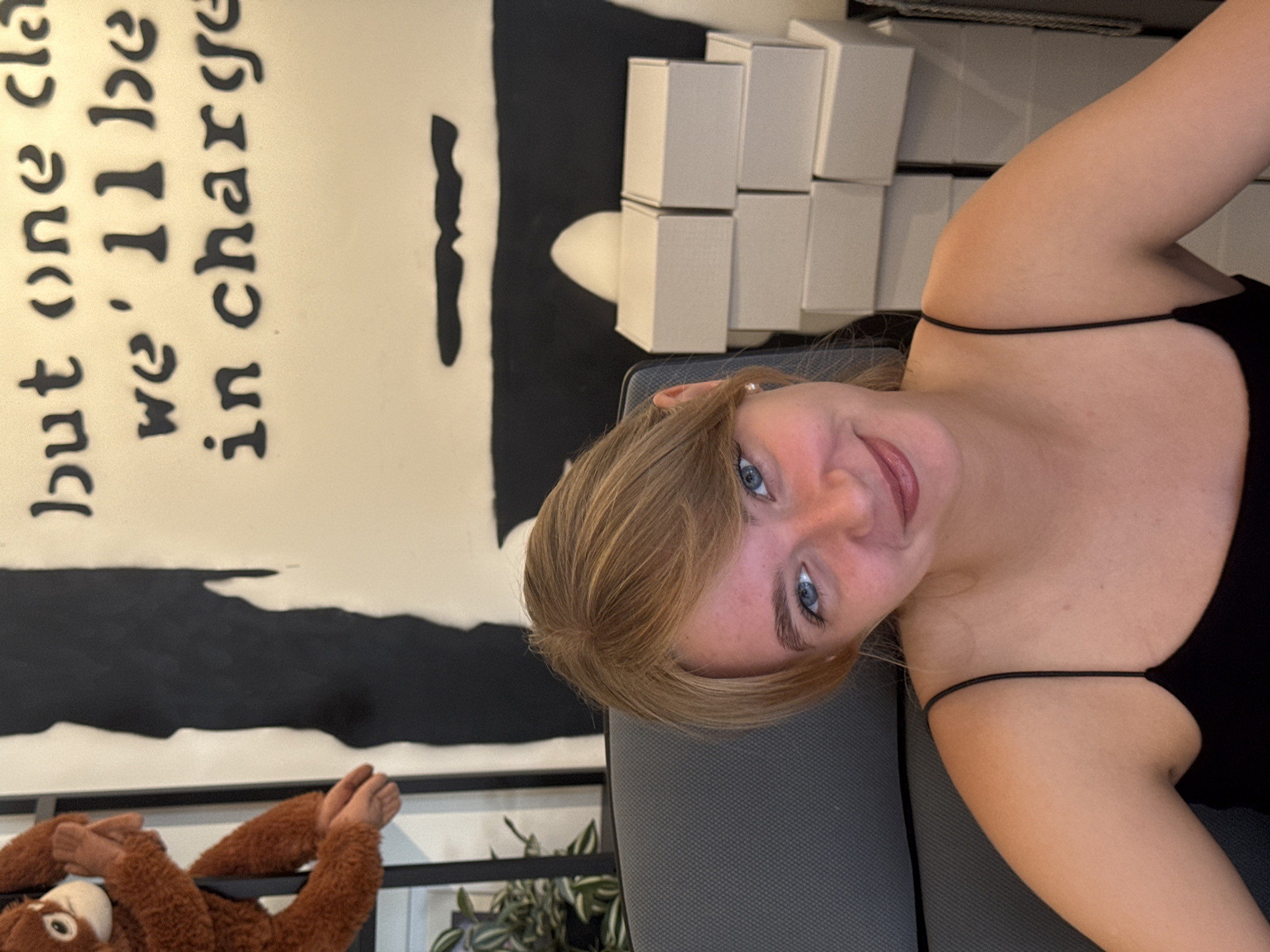 A young woman with blonde hair and blue eyes is seated in a chair, looking at the camera with a slight smile. She is wearing a black spaghetti strap top. Behind her, a wall features black lettering that reads "but one cla we'll be in charge." To her left, a stuffed monkey toy hangs from a metal structure, and to her right, a stack of small white boxes is visible. The scene appears to be indoors, possibly in a room or an office setting. The specific location within the city of Kremenchuk, Ukraine, cannot be determined from the visual information provided in the image.