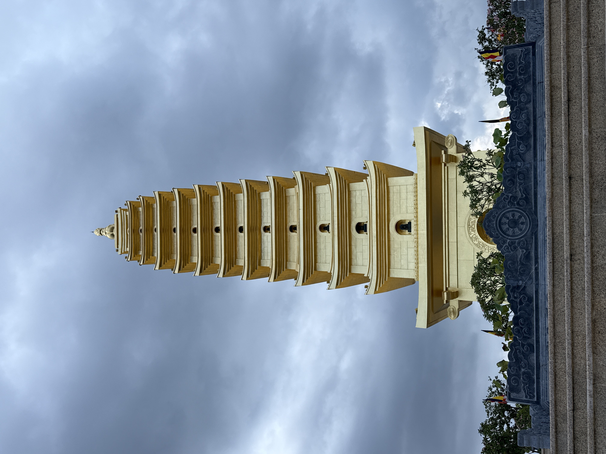 A vertical shot shows a yellow pagoda reaching into a cloudy sky in Diên Khánh, Vietnam. The pagoda, composed of multiple tiered sections, appears to be made of a light-colored stone or concrete that is coated in gold. It is intricately designed, with a number of identical tiers decreasing in size as they rise to a rounded pinnacle. On the right edge of the frame, a section of a dark gray stone wall with carved designs is visible, adorned with leafy plants. The sky is overcast with gray clouds, providing a stark contrast to the golden pagoda.