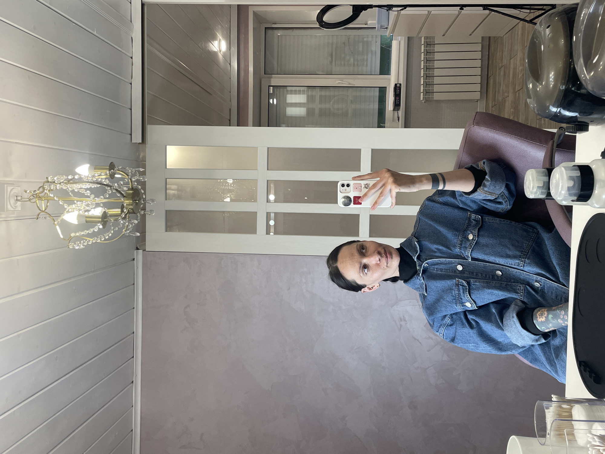 A woman is taking a mirror selfie in what appears to be a hair salon or beauty studio. The image captures her reflection, holding a white smartphone with stickers on the back. She is seen from the chest up, with dark hair pulled back, wearing a black top underneath a blue denim jacket. Notable personal details include a black band tattoo on her left arm, a colorful floral tattoo on her right forearm, and piercings on her nose and ears. Her expression is neutral as she looks towards the phone.
The setting reveals elements typical of a salon. To the right, a dark-colored salon chair is partially visible, with two clear, dome-shaped hair processors or dryers suspended above it. On a counter below, several white and clear plastic bottles and a black mat suggest hair products or tools. A ring light is reflected in the upper right, indicating professional lighting.
The interior design features a light-colored, horizontally paneled wall and ceiling on the left. The wall behind the woman has a textured, light purple or grey finish, possibly Venetian plaster. A white door with multiple rectangular glass panels is prominent in the background, reflecting an exterior window and a white radiator, suggesting it's an indoor commercial space. A decorative chandelier with crystal-like elements and multiple lights hangs from the paneled ceiling on the left. No discernible text is visible in the image. The time of day appears to be daytime, enhanced by artificial lighting.