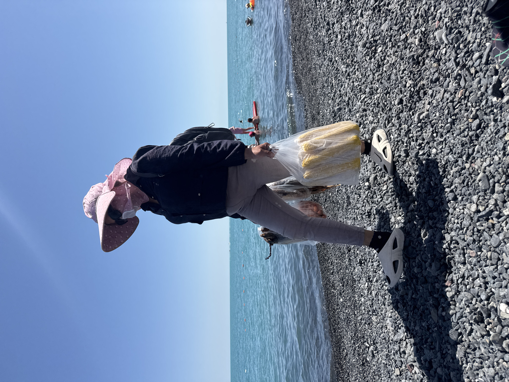 A person wearing a pink sun hat and a dark jacket walks along a rocky beach next to a body of turquoise water. They are carrying a clear plastic bag filled with corn on the cob and another bag with fish hanging from it. The person is also wearing gray pants, white clogs, and black socks. In the background, several people are visible in the water, some on a red float. The sky is clear and blue. The scene appears to be a public beach on a sunny day. The location is specified as the city of Ақши, Kazakhstan.