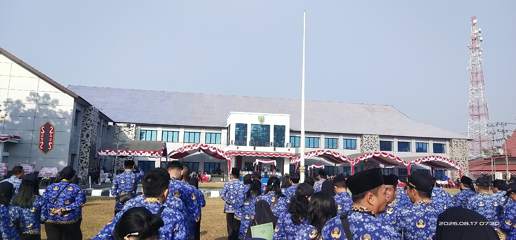 This image captures an outdoor gathering in front of a large, modern building in Tamiang Layang, Indonesia. The building appears to be a government office, possibly a municipal building, with its name "KANTOR..." visible on the facade. A tall flagpole stands prominently in the foreground, and the scene is set against a clear, bright sky.
A crowd of people, predominantly men and women in matching blue uniforms with a recurring golden emblem, are assembled on a grassy field in front of the building. The uniforms feature a repeating pattern, and many of the men are wearing black peci hats. They appear to be participating in an official event or ceremony, as indicated by the presence of red and white draped awnings and a stage-like area within the building's entrance.
The architectural style of the building is modern, with multiple floors, large windows, and decorative patterned panels on its exterior. A red and white striped antenna tower is visible in the background, suggesting the presence of communication infrastructure. The overall atmosphere suggests a formal or civic occasion taking place in a public space.