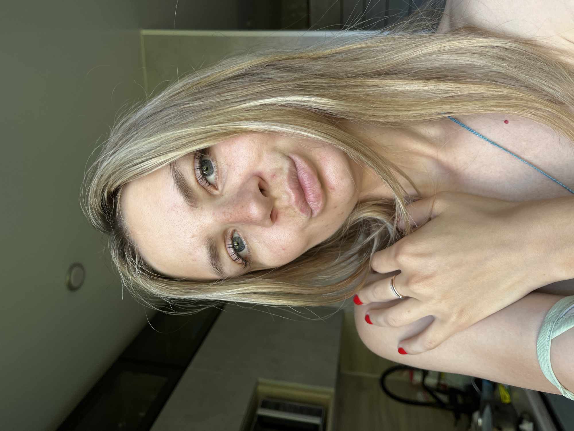 A blonde woman is captured in a selfie, holding her arm across her chest with her right hand. She has blue eyes, freckles, and is wearing red nail polish. A delicate ring adorns her ring finger. The background suggests an indoor setting with a window or door to the right, revealing a blurred view of what appears to be an urban environment, possibly a kitchen or living area with some cabinetry visible on the left. The lighting is natural, coming from the right side, casting a soft glow on the woman's face. There is no indication of the specific location in the city of Chaikovsky, Russia.