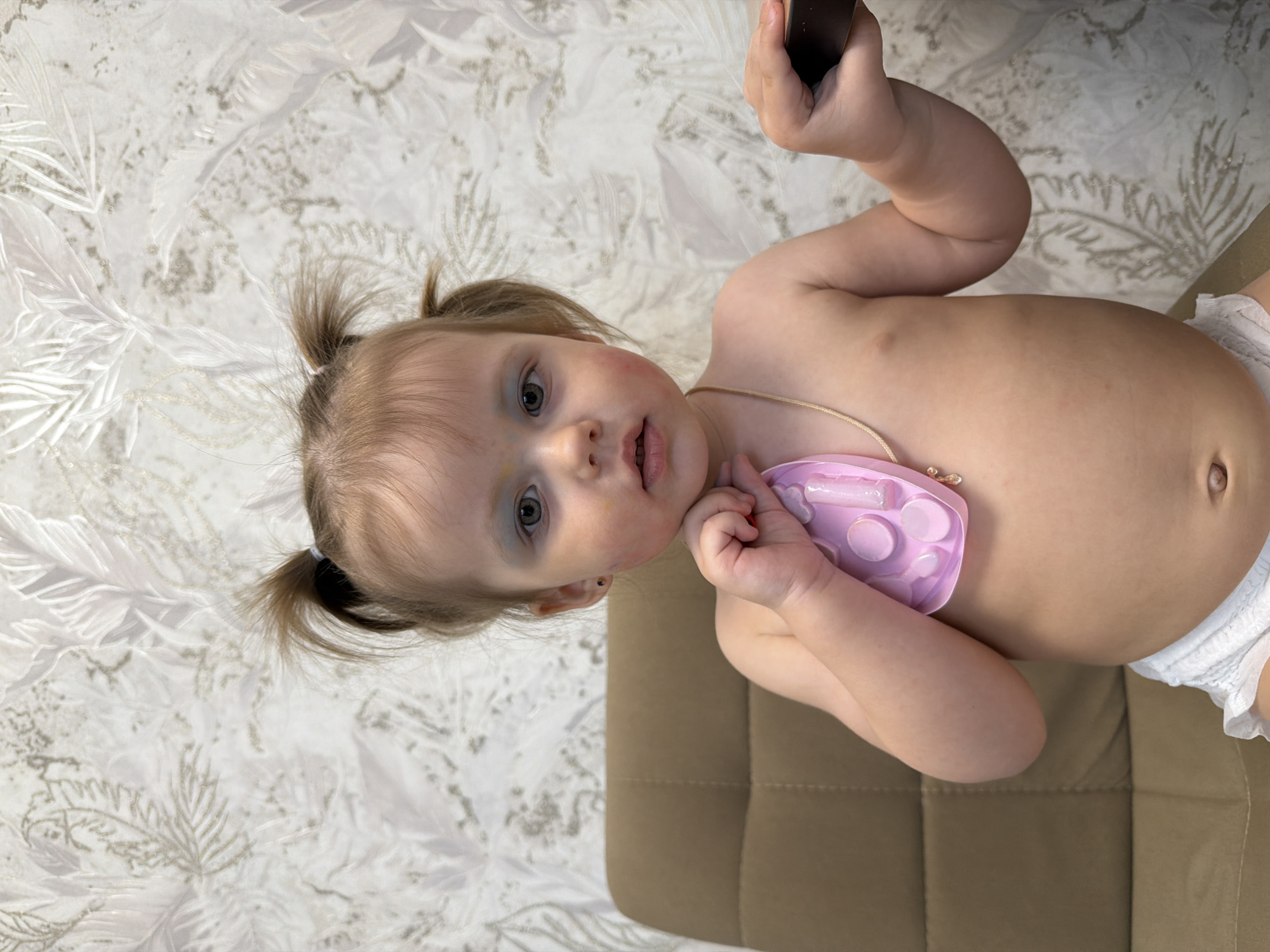 A fair-skinned toddler with blonde pigtails is the central subject of the image. The toddler has light eyes and appears to have some redness or marks on their face. The child is mostly undressed, wearing what appears to be a diaper and a light pink necklace with a plastic component. The child holds a black object in their right hand, raised above their head. The child's left hand is near their face.
The toddler is positioned on a light brown textured surface, possibly a chair or sofa. The background consists of a patterned wallpaper with a repeating design. The location is not possible to determine from this image alone.