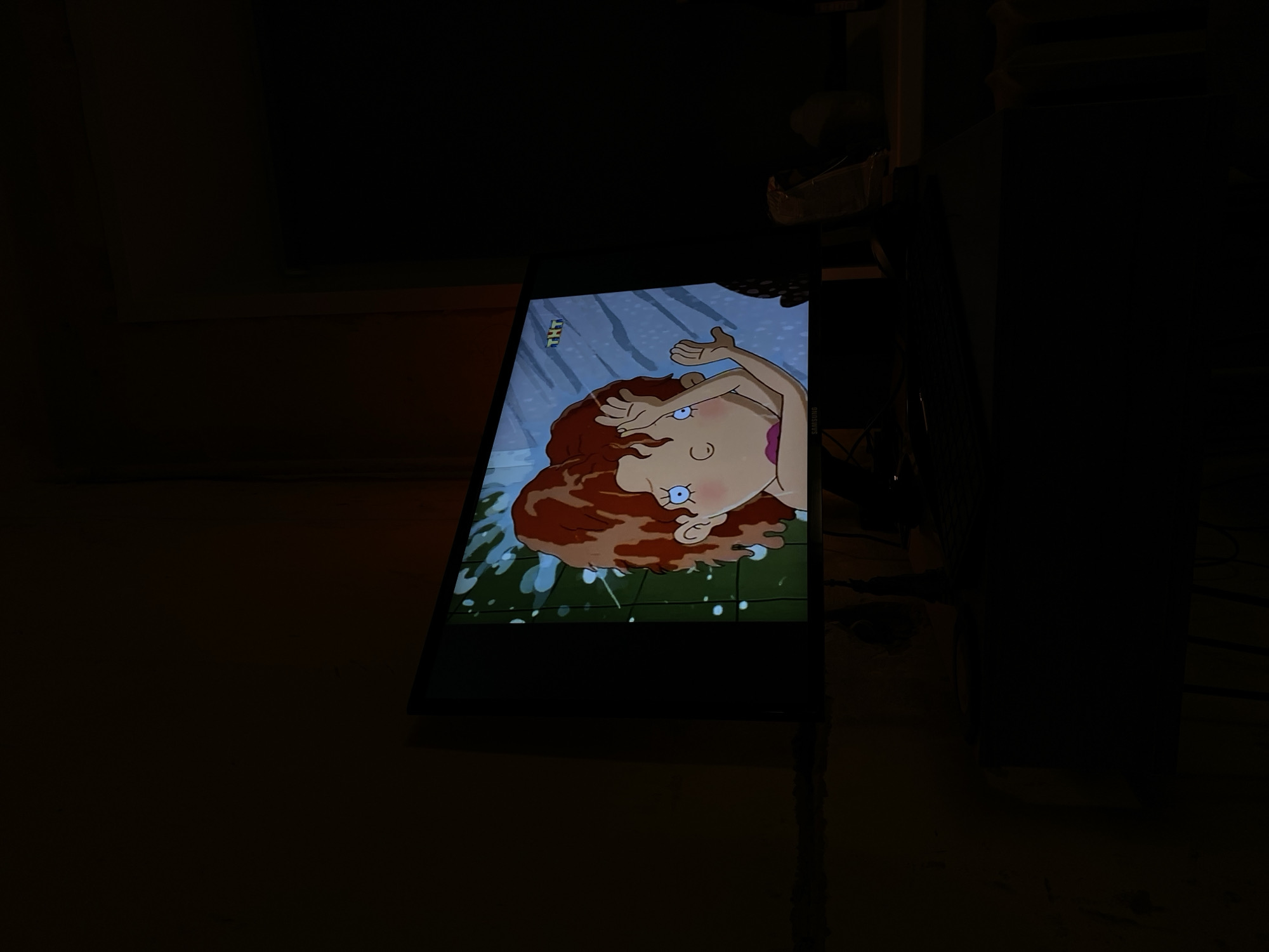 The image shows a television screen displaying a cartoon scene of a red-haired character. The screen is surrounded by darkness, with the corner of a wall or furniture visible on the left and a dark object resembling a cabinet or shelf on the right. The scene appears to be indoors, possibly a living room or bedroom. The location is unknown and cannot be determined as Votkinsk, Russia.