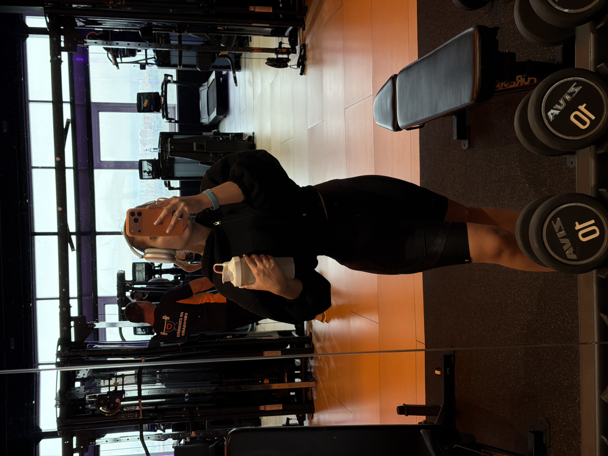 A mirror selfie of a woman holding a water bottle and wearing wireless headphones in a gym. The gym has large windows overlooking a city. Based on the visible landmarks and architectural style, the city is likely Krasnoyarsk, Russia. The gym equipment includes weight machines, a bench, and dumbbell rack with Avis branded weights. Another person is partially visible in the background.