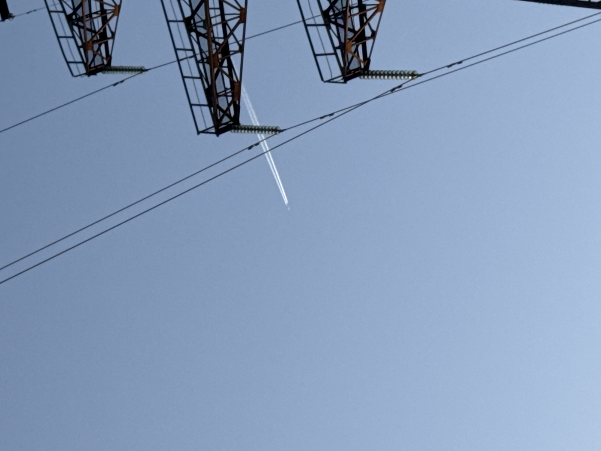The image shows a low-angle view of electrical transmission towers against a clear, light blue sky. Two parallel transmission lines run diagonally across the frame. An airplane with a visible contrail is ascending through the center of the image, passing behind the electrical towers and wires. The electrical towers are dark, rusty brown, and appear to be made of latticed metal. Insulators connect the wires to the towers. The location is Chaikovsky, Russia.