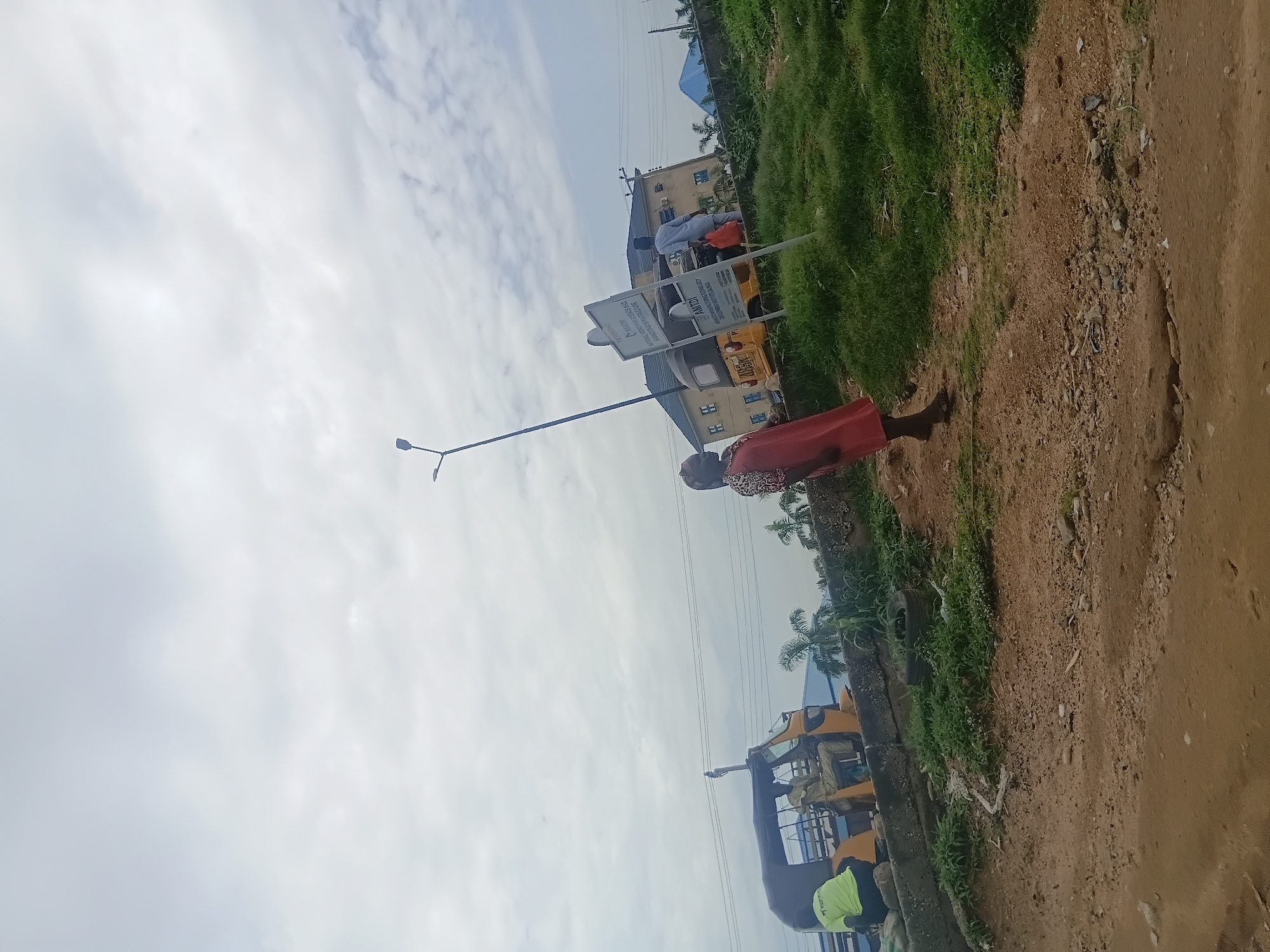 The image depicts a street scene in Jalingo, Nigeria. The main subjects are a few people, one woman in the foreground wearing a red dress and a patterned top, and several individuals near a yellow motorized tricycle (a common mode of transport in Nigeria, often called an "okada" or "keke"). The scene also includes buildings in the background, a grassy embankment with some debris, and a dirt road.
The activity appears to be casual, with people gathered near the tricycle, possibly waiting for a ride or engaged in conversation. The woman in the foreground is standing and looking towards the right of the frame.
Notable details include a lamppost with two lights, signs on posts near the tricycle, and power lines running across the sky. The weather seems to be overcast with clouds filling the sky. The time of day is difficult to determine precisely, but the lighting suggests it is daytime. The presence of the motorized tricycle and the style of the buildings are strong location cues for Nigeria.
There is visible text on the signs near the tricycle. One sign appears to be white with black lettering, and another is yellow with black text. While the text is not fully legible, one sign has "ANTED" clearly visible.
The overall impression is of a typical street scene in a Nigerian town on a cloudy day.