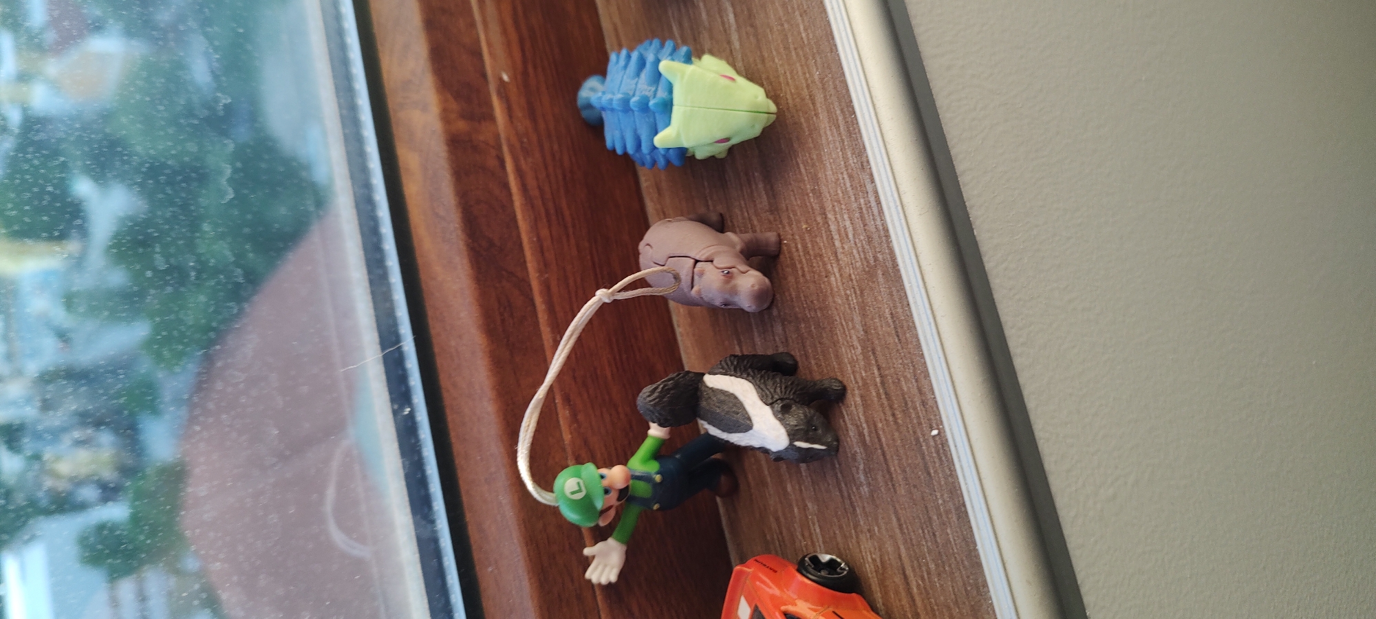 A still-life photograph shows a collection of toy figures arranged on a brown, wood-grained surface. From the top, there is a blue and lime green spiky toy, a light brown hippo figure with a white string attached, a black and white animal figure resembling a tapir, a Luigi figurine with green hat and blue overalls, and a partially visible orange toy car at the bottom. The toys are positioned along the edge of a window frame. A window is visible on the left, revealing a blurred outdoor scene. The scene is lit by natural light coming from the window. The backdrop on the right is a light gray wall.