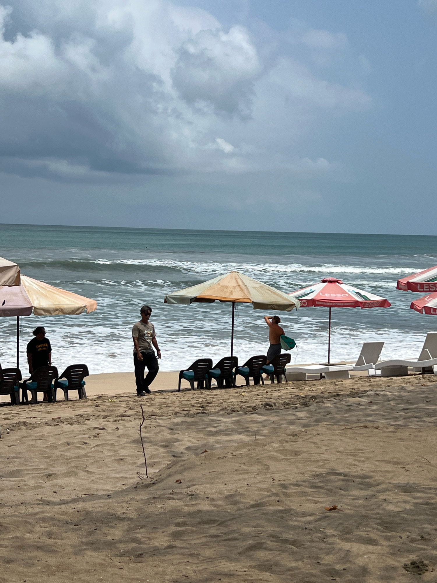 A wide shot captures a beach scene in Kuta, Indonesia. The sky is overcast with heavy, dark clouds, and the sea is choppy with white-capped waves. In the foreground, a sandy beach is lined with beach chairs and umbrellas. Three people are visible on the beach. One person stands near a beach umbrella, looking out at the sea. Another person walks across the sand, and a third person is further in the background, holding a surfboard. Beach umbrellas are set up, some with chairs positioned beneath them, suggesting a recreational area. The overall atmosphere appears to be that of a beach on a cloudy day, with a few people present.