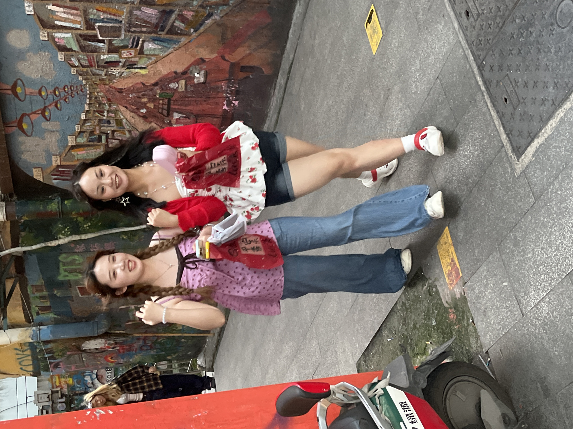 Two young women are posing for a photo in Jiulongpo District, China, standing on a city sidewalk alongside a mural-covered wall. One woman wears a white top with red floral prints, a red cardigan, and dark denim shorts, while the other sports a purple polka-dot top and blue jeans. They are holding identical red promotional items. A scooter partially occupies the foreground of the image, and a man in a checkered shirt stands behind them. The setting includes street art, a marked pedestrian path, and a manhole cover.