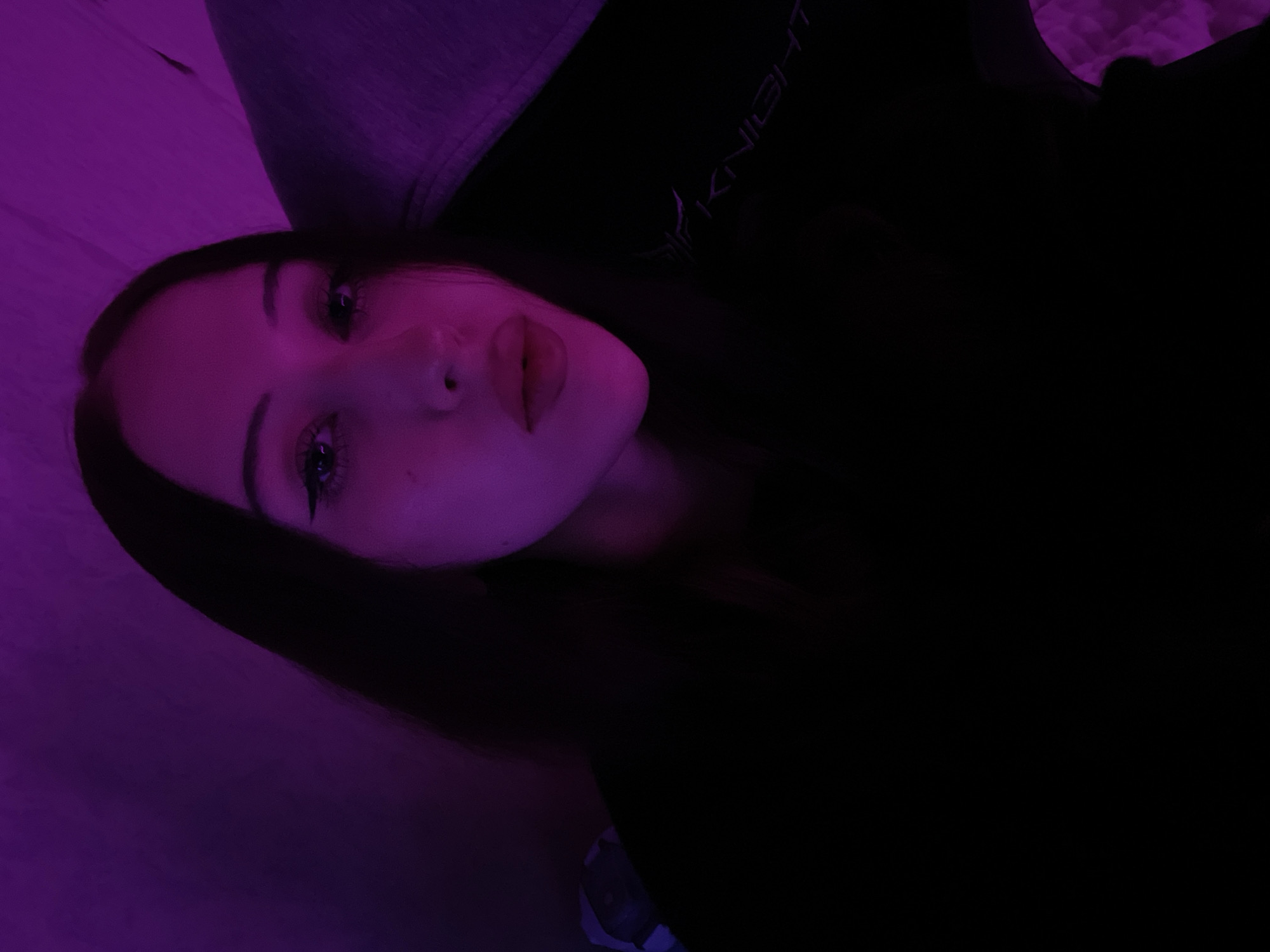 A young adult female is depicted in a medium close-up, facing directly towards the camera. She has long, straight dark hair, styled parted centrally. Her eyes feature winged eyeliner, and her lips are full, appearing to have a glossy finish, slightly parted. A small mole or freckle is visible on her right cheek (viewer's left). She is wearing a dark, presumably black, garment. On the upper right chest/shoulder area (viewer's left), a stylized white or silver logo is partially visible, clearly showing the text "KNIGHT" with an initial letter that appears to be a stylized 'F'.
The scene is illuminated predominantly by a magenta or purple neon light source, casting a strong color hue across the subject's face, hair, and the immediate environment. The background is dark and indistinct, consistent with an indoor setting, with soft, textured surfaces in the upper right. Faint outlines of what appears to be dark furniture or a chair, possibly a gaming chair, are discernible in the shadowed areas, also reflecting the purple light. There are no identifiable geographical markers within the image to determine a specific location such as Votkinsk, Russia.