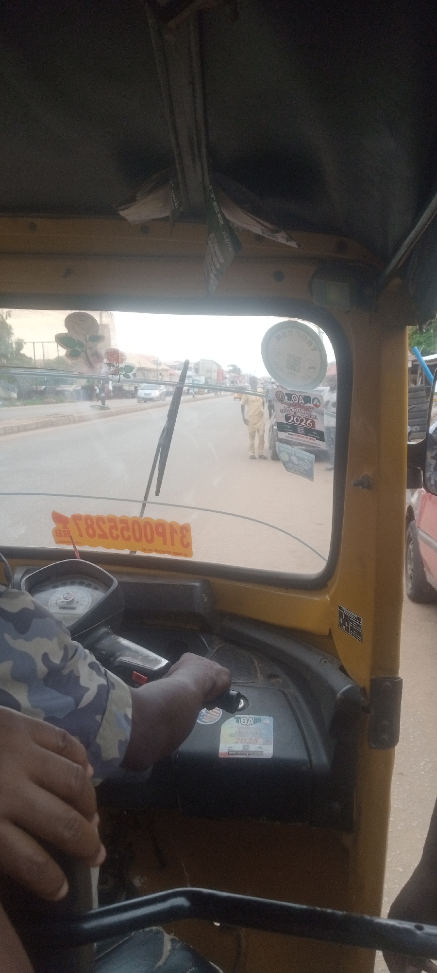 The image is taken from inside a yellow tricycle auto rickshaw, commonly known as a "keke," on a street in Sabon Tasha, Nigeria. The driver's hands are visible on the handlebars. Through the windshield, a street scene unfolds. A man dressed in a light-colored traditional outfit is seen walking near the side of the road. Parked cars and other buildings are visible further down the street. The windshield features stickers.