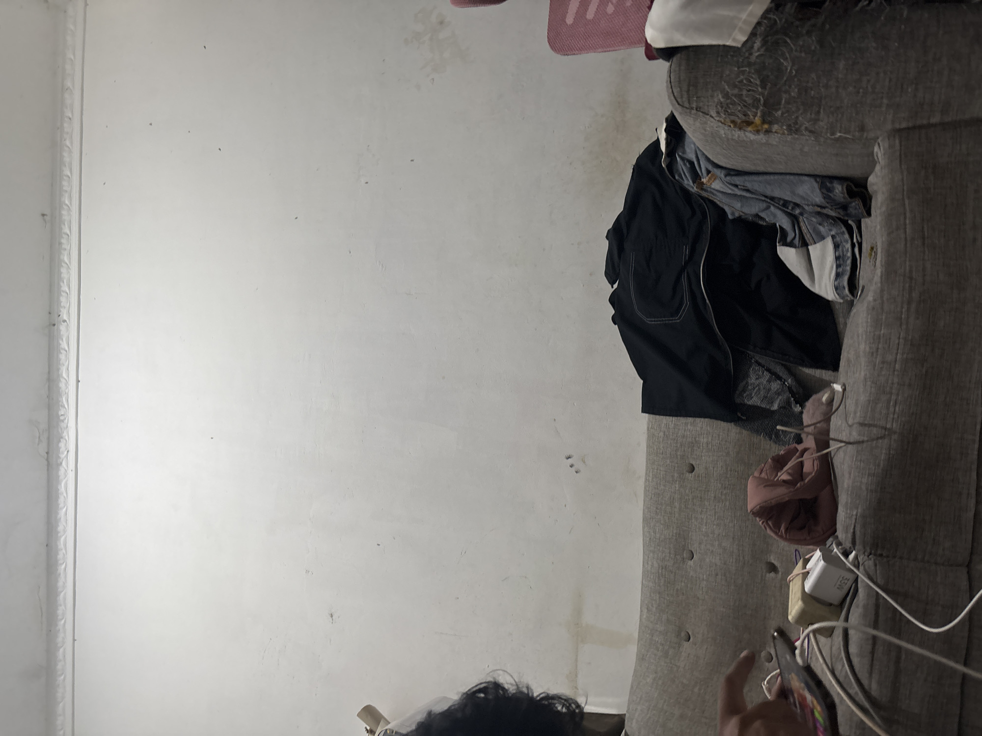 The image depicts a dimly lit indoor scene, likely a living room, in North Jakarta, Indonesia. A person's hand is visible on the left side of the frame, interacting with a smartphone that is connected to a charging block and multiple cables. The smartphone is placed on a grey sofa, which is also covered with various items of clothing including a black jacket, a pink garment, and a denim jacket. The sofa appears worn, with visible damage to its upholstery on the right side. In the background, a white wall with a decorative molding at the top is visible. A pink chair is partially visible on the far right of the image.