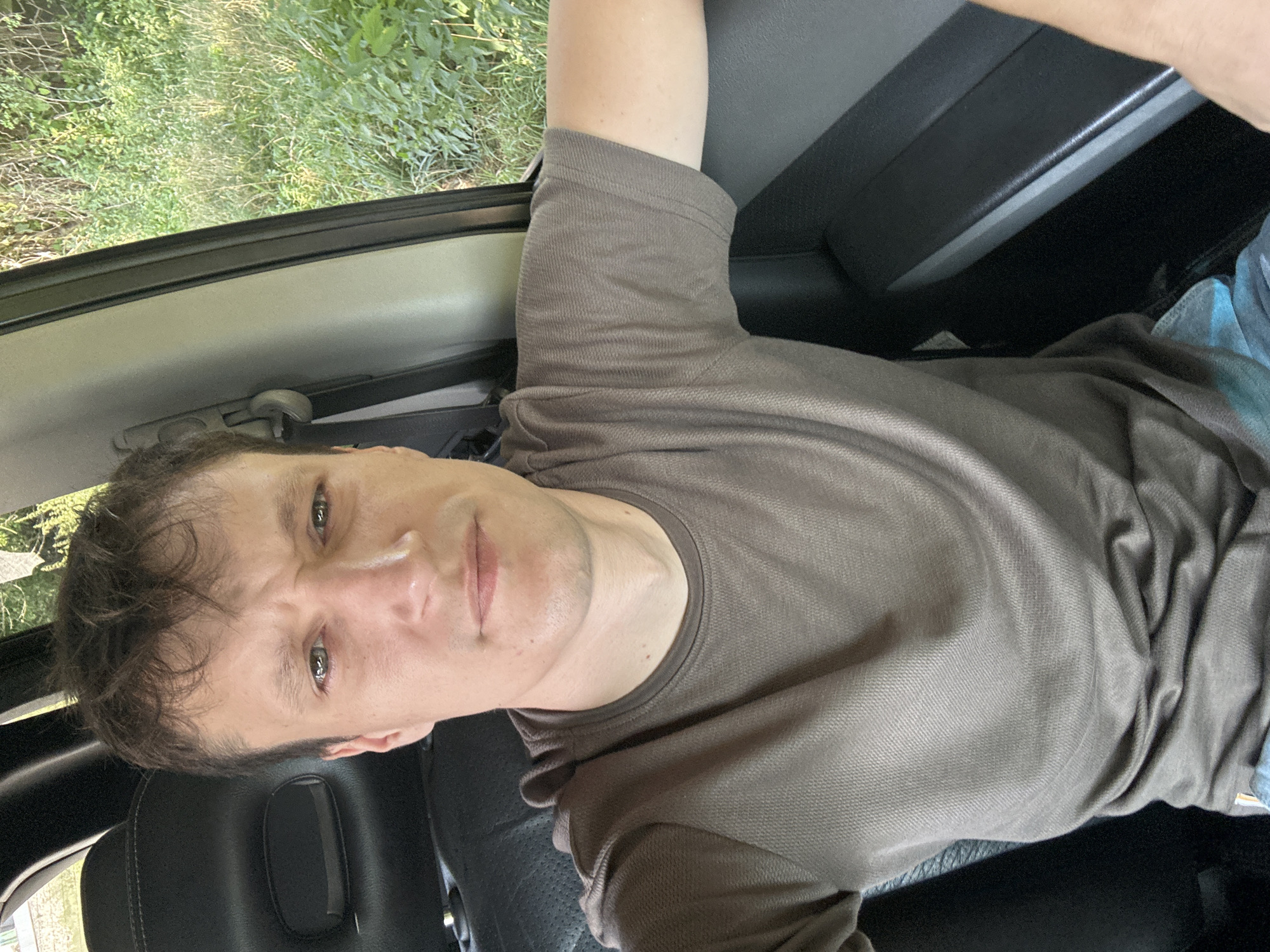 A man with short, dark, wavy hair and fair skin is sitting in the driver's seat of a vehicle, taking a selfie. He is wearing a plain, short-sleeved, dark brown t-shirt. His facial expression is one of mild annoyance or contemplation, with his brow furrowed and his mouth slightly downturned. The background shows the interior of the car, including a black leather headrest and seat, a gray interior door panel, and a portion of the side window revealing lush green foliage and trees outside. The image appears to be taken during daylight. The location is identified as Makiv, Ukraine.