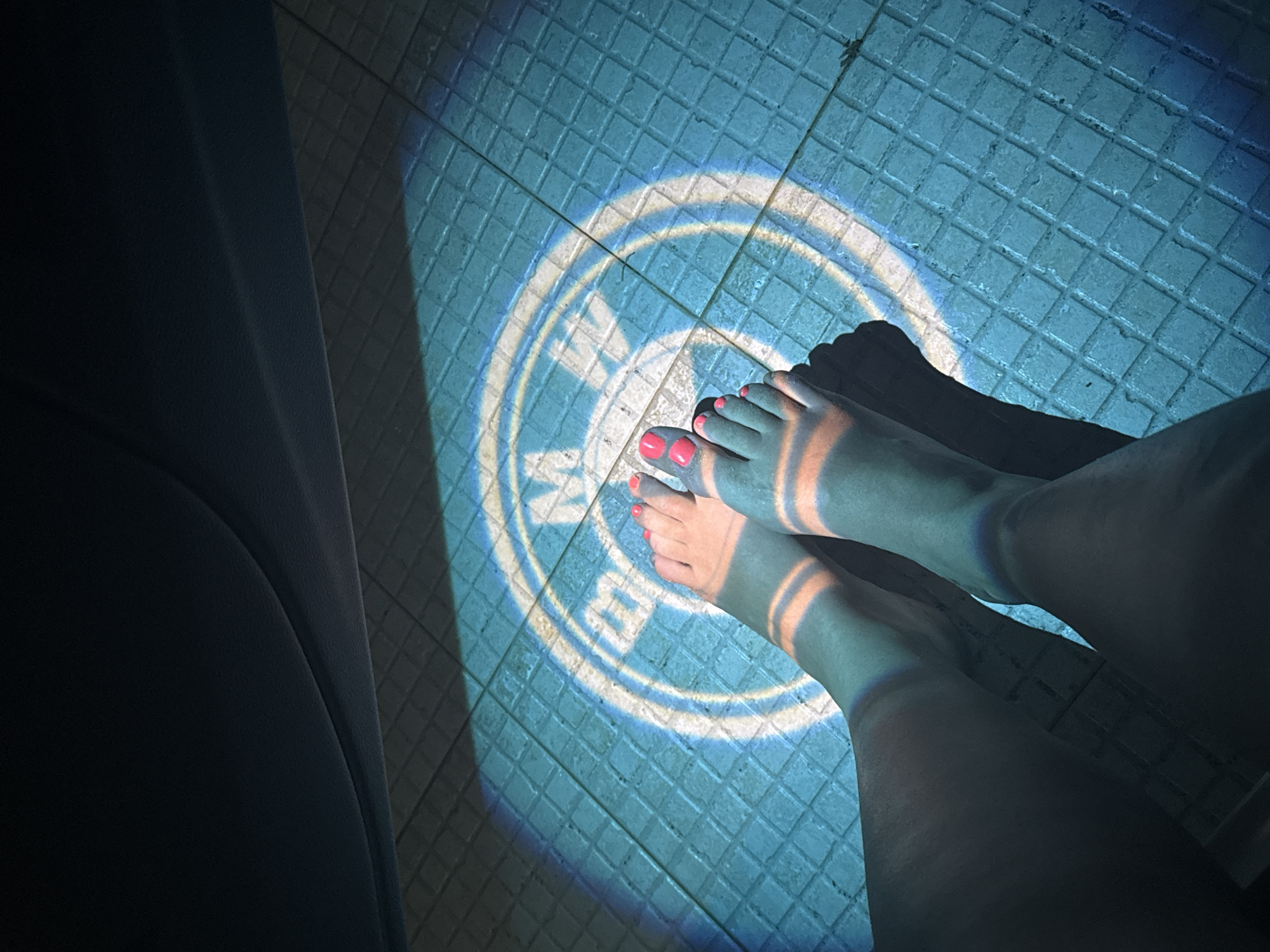 In an outdoor, tiled setting, a person's feet, with bright pink toenail polish, are illuminated by a projected BMW logo light. The logo is displayed on the floor, casting a blue and white glow that highlights the texture of the tiles. Shadows from the car or an overhang fall across the scene, adding depth and contrast to the image. The overall ambiance suggests a nighttime or low-light environment, possibly in a parking area or a similar urban outdoor space.