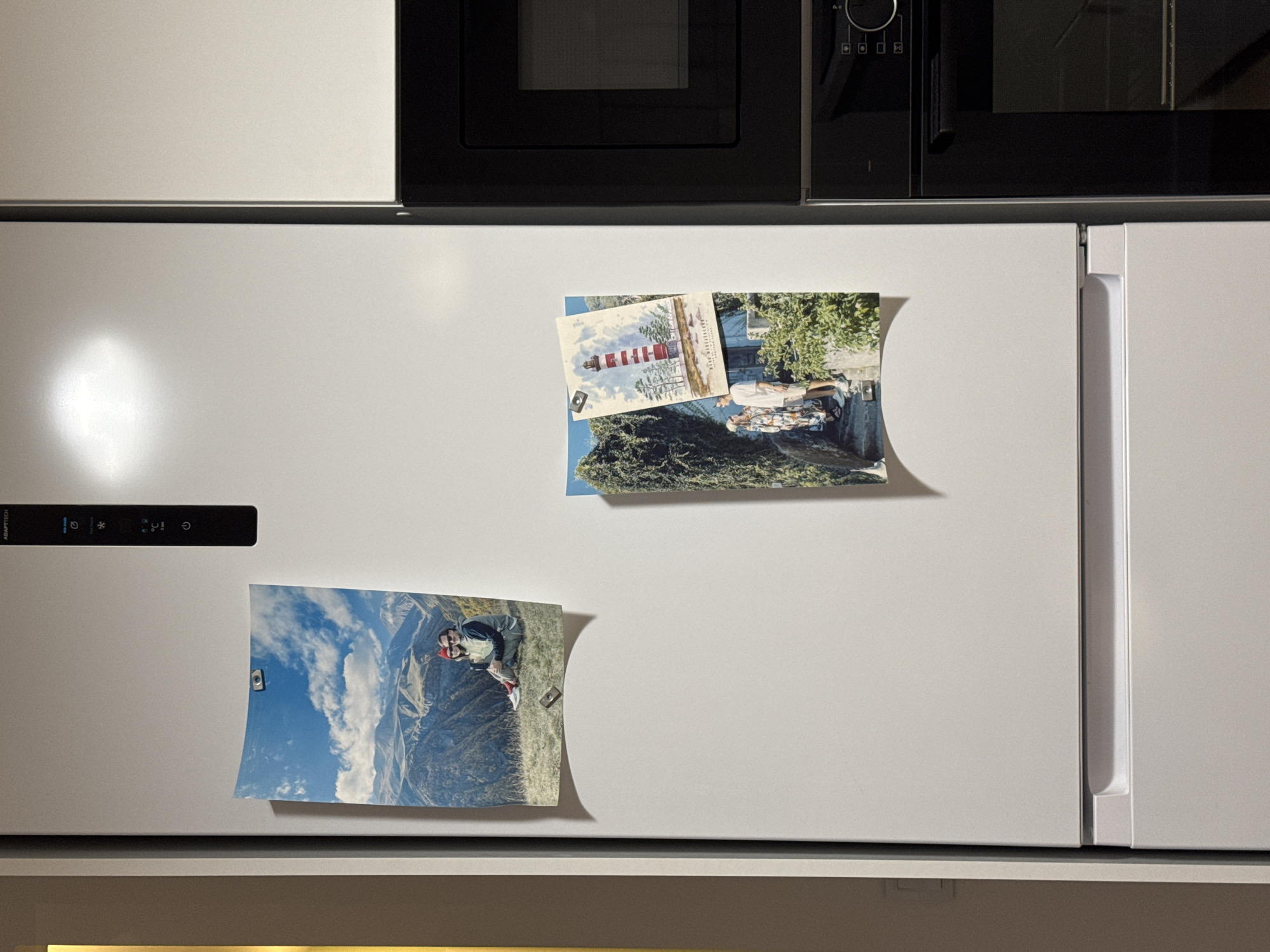 The image displays a domestic kitchen setting, likely in Izhevsk, Russia, featuring white built-in appliances. The central focus is a large white refrigerator, positioned beneath two black built-in ovens or microwaves.
The refrigerator's left door has a dark control panel with illuminated symbols indicating functions like temperature control (snowflake), air circulation (fan), and a power button. The brand name "ADAPTITECH" is partially visible on this panel. Two mementos are affixed to the refrigerator door with small, rectangular magnets.
The upper memento is a postcard depicting a red and white striped lighthouse surrounded by trees, with some text in Cyrillic script partially visible below it, which appears to say "БИРЮЧИЙ ЛОСОСЬ" (Biryuchy Salmon), possibly referencing a location or theme. Below the postcard is a photograph showing two people walking on a path near dense green foliage and a building, suggesting an outdoor travel scene.
The lower memento is a vertical photograph or postcard featuring a person, likely a man, wearing a red cap, dark jacket, and light-colored pants, sitting on a grassy hillside. The background reveals a vast mountainous landscape under a cloudy sky.
The black appliances above the fridge are sleek and modern, with one showing a control knob and several square buttons. The overall scene is static, capturing a moment in a household setting decorated with personal travel memories. The lighting appears to be indoor, with a bright reflection on the left side of the refrigerator surface.