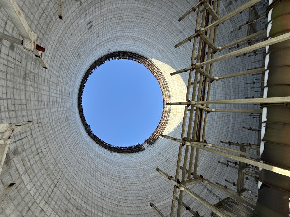 The image presents a striking upward-looking perspective from within a colossal, circular industrial structure, strongly resembling a cooling tower. The vantage point is directly from the base, emphasizing the immense height and scale of the edifice.
The interior walls of the structure are constructed from light grey concrete, characterized by a grid-like or waffle pattern composed of numerous square or rectangular panels. This textured surface appears weathered and somewhat aged, with subtle variations in tone suggesting exposure to elements over time. The cylindrical form of the tower tapers slightly towards the top, perfectly framing a pristine, bright blue sky in a distinct circular opening directly overhead. The clarity of the sky indicates fair weather and daytime.
On the right side of the image, a complex internal framework or scaffolding system is visible, extending vertically. This structure is composed of numerous light-colored beams, possibly made of wood or a similar material, interconnected by horizontal cross-members, forming a multi-tiered, angular pattern. Further to the right and slightly out of focus, darker, rectangular or trapezoidal panels appear to be attached to or supported by this framework. These elements are likely components of the cooling tower's fill material, designed to increase the surface area for water evaporation.
The lighting within the tower is natural, emanating from the circular opening at the top. The direct sunlight highlights portions of the right wall and the internal framework, creating areas of brightness and casting subtle shadows that add depth to the massive structure. There are no people visible in the image, nor is any discernible text present. The overall impression is one of vast industrial architecture, perhaps abandoned or under construction, with a dramatic interplay of concrete, structural elements, and open sky.