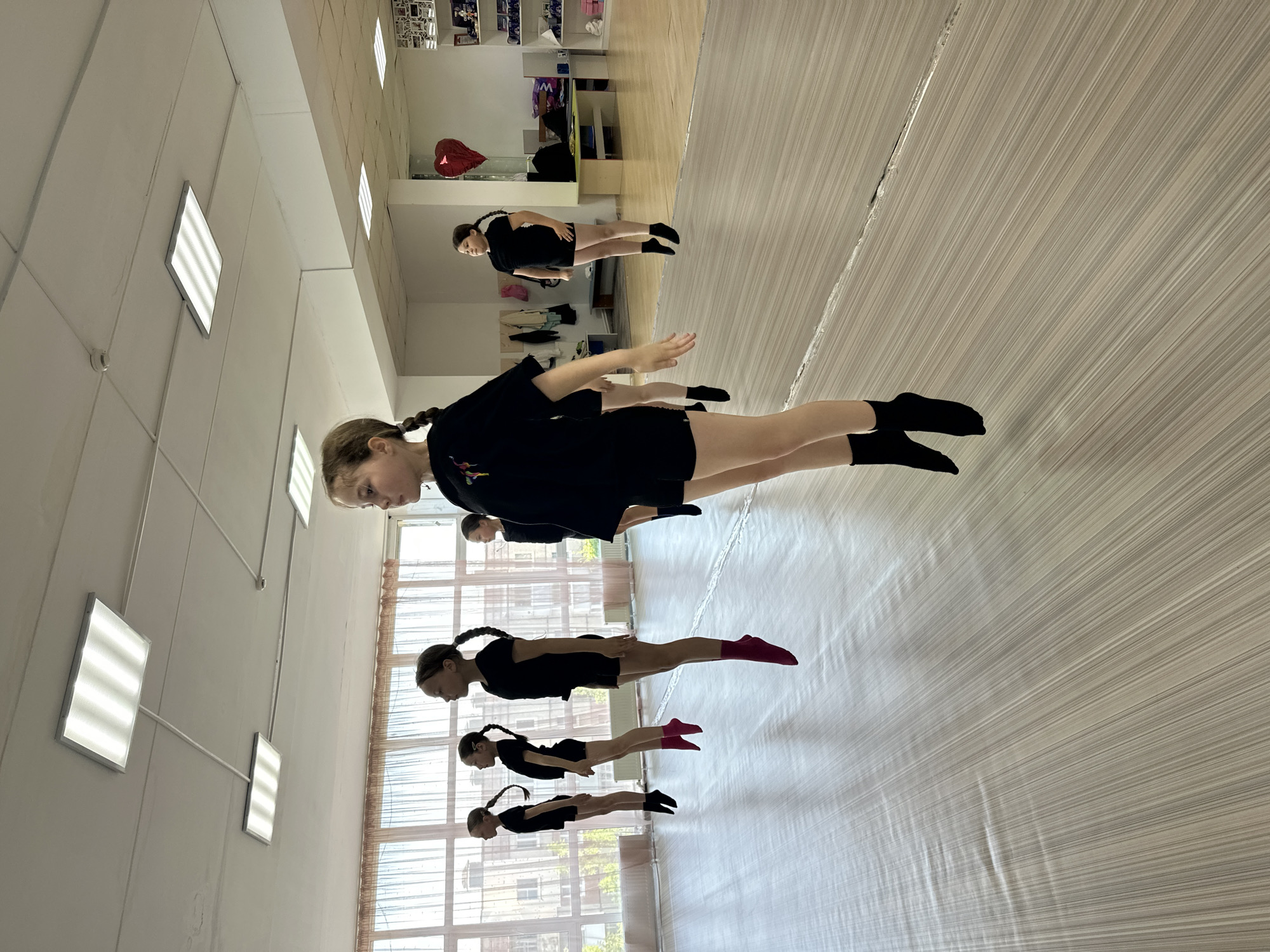 A dance studio in Votkinsk, Russia. A line of five young girls with braided hair, wearing black outfits and socks (some pink), are practicing a dance routine in a dance studio. They are arranged in a line facing the same direction. The studio has a light-colored wooden floor and large windows on one side and a mirror on the opposite wall. The room is lit by fluorescent ceiling lights. There is no other distinct feature.