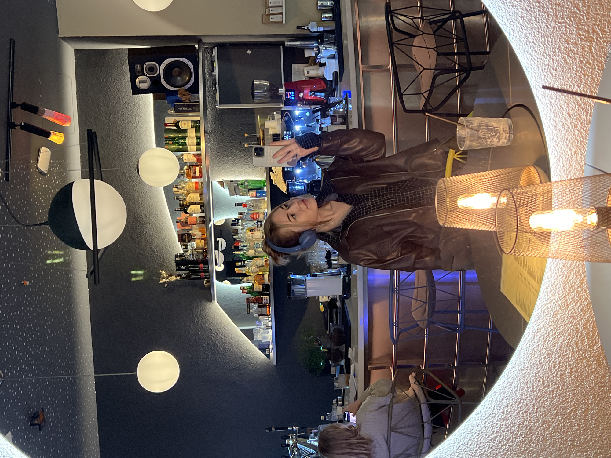 The image depicts the interior of a contemporary bar or restaurant located in Rafailovići, Montenegro, captured as a mirror selfie.
In the foreground, a young Caucasian female with light brown hair, wearing blue over-ear headphones, a brown leather jacket, and a dark plaid shirt, takes a selfie using a light-colored smartphone held in her right hand. She is positioned centrally in the mirror's reflection, looking towards the phone. On a dark circular table reflected in the lower portion of the mirror, two mesh-patterned table lamps with exposed filament bulbs illuminate a clear glass with residual contents and a stirring stick, alongside a yellow menu or document. The mirror itself features a wide, textured, light-toned frame.
The background, also reflected, shows a well-stocked bar area. Multiple shelves display various liquor bottles, illuminated by strip lighting. The bar counter features professional coffee machines (one red, one silver), grinders, and an assortment of bar tools and glassware. To the left, a second Caucasian female with blonde hair, wearing a light-colored long-sleeved top, is seated at the bar, facing away from the camera, appearing to interact with a device or menu. Above the bar, a large black speaker is visible on a shelf. The walls are dark with a textured finish, and the ceiling is dark with numerous small white dots, simulating a starry effect. Various modern light fixtures include spherical white pendant lights, a large dark and white spherical fixture, and track lighting with tubular multi-colored bulbs. Modern wire-frame chairs, both black and light blue, are visible around the seating areas.