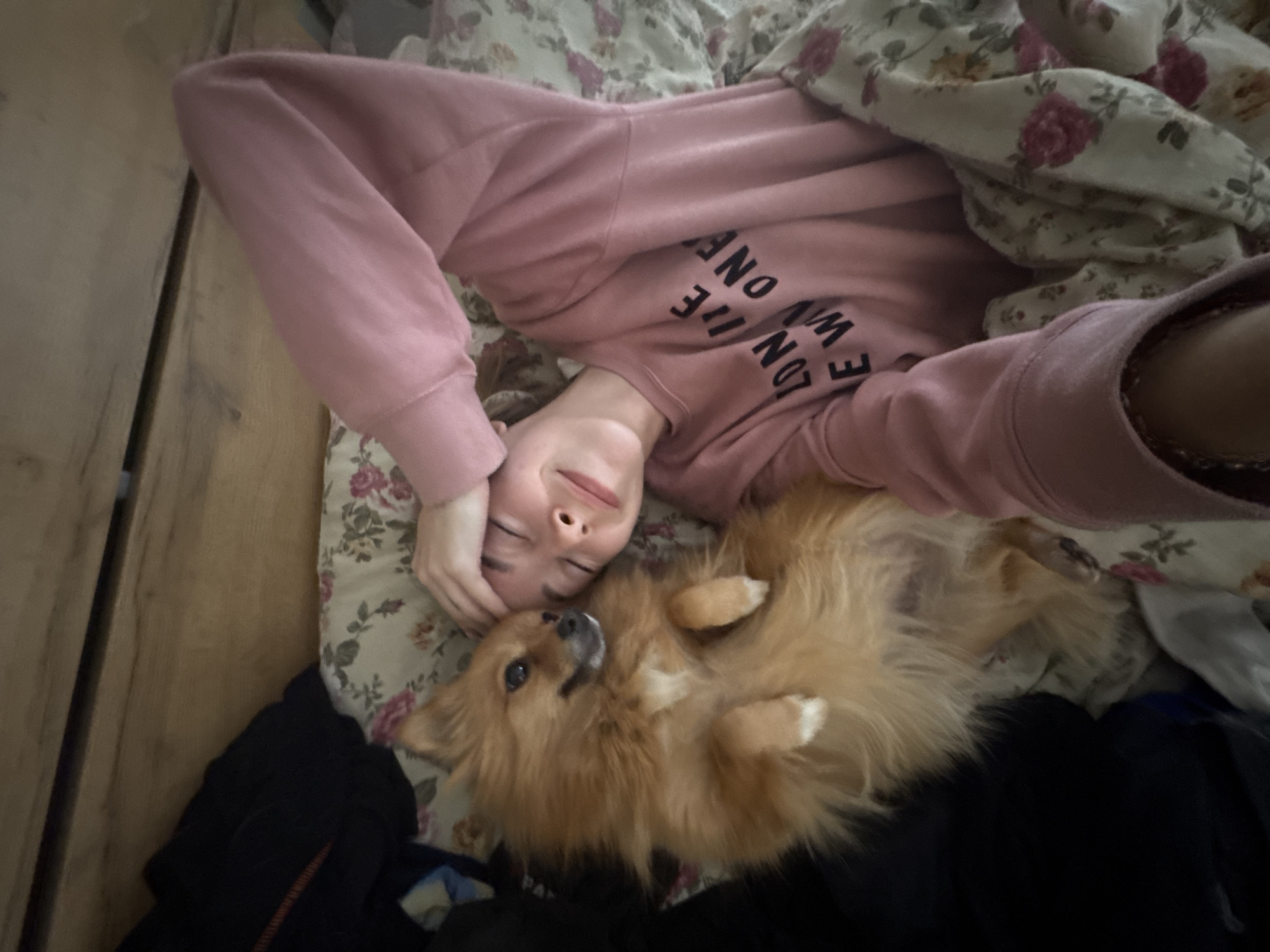 A young person in a pink sweatshirt is lying on a floral patterned bed under a floral blanket with a Pomeranian dog. The dog is lying on its back with its paws up. The sweatshirt says “LONDON ONE WE.” Some dark clothing is next to the bed on a wooden floor. The location is not identifiable from the image.