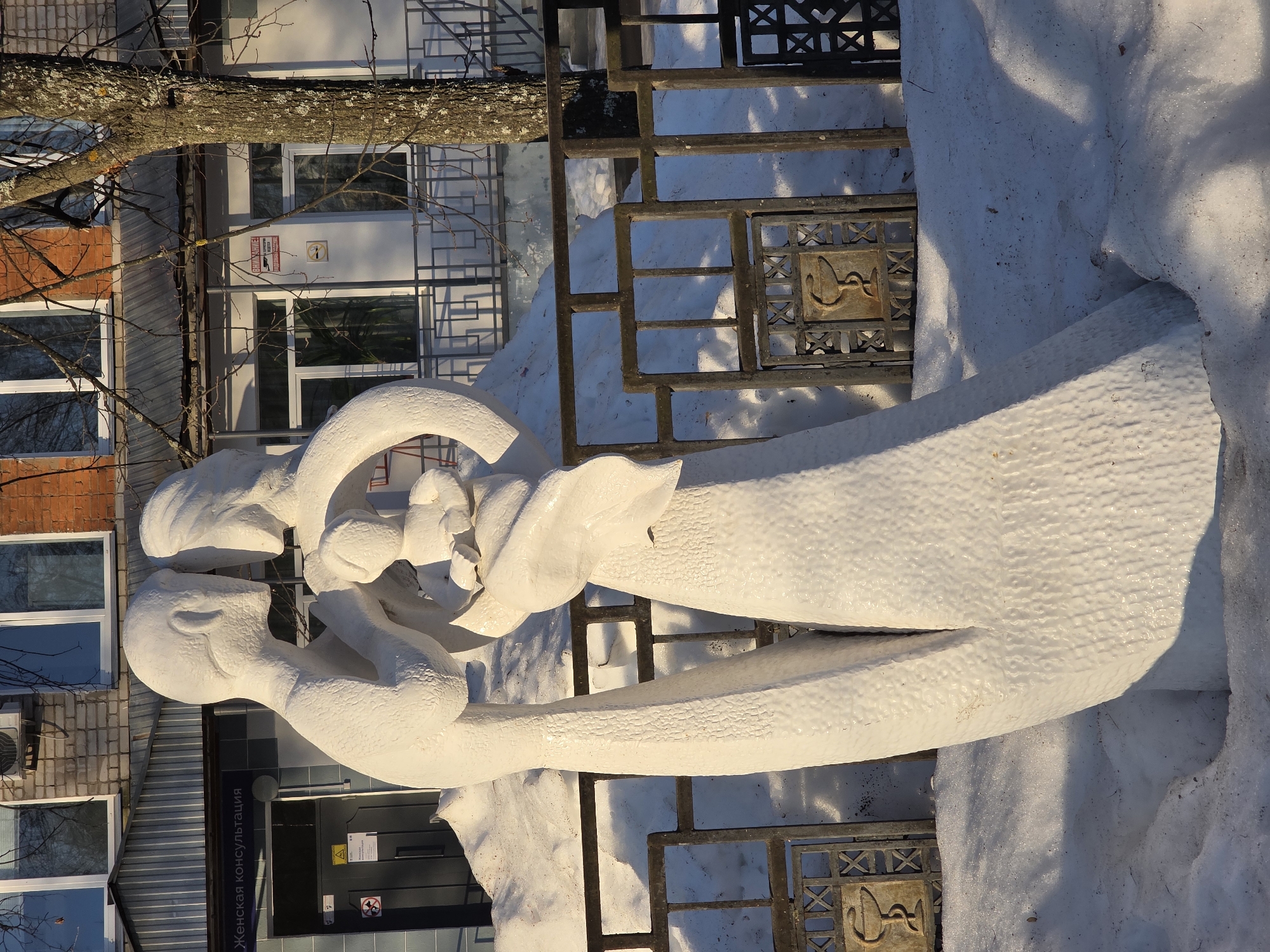 The image shows a white sculpture of a mother and child located outside the "Zhenskaya Konsultatsiya" (Women's Consultation) clinic in Chaikovsky, Russia. The sculpture depicts two figures embracing, covered with snow. It is situated next to a metal fence with decorative panels featuring medical symbols. The background includes the clinic's building and a tree. The scene is likely taken during winter, as evident by the presence of snow.