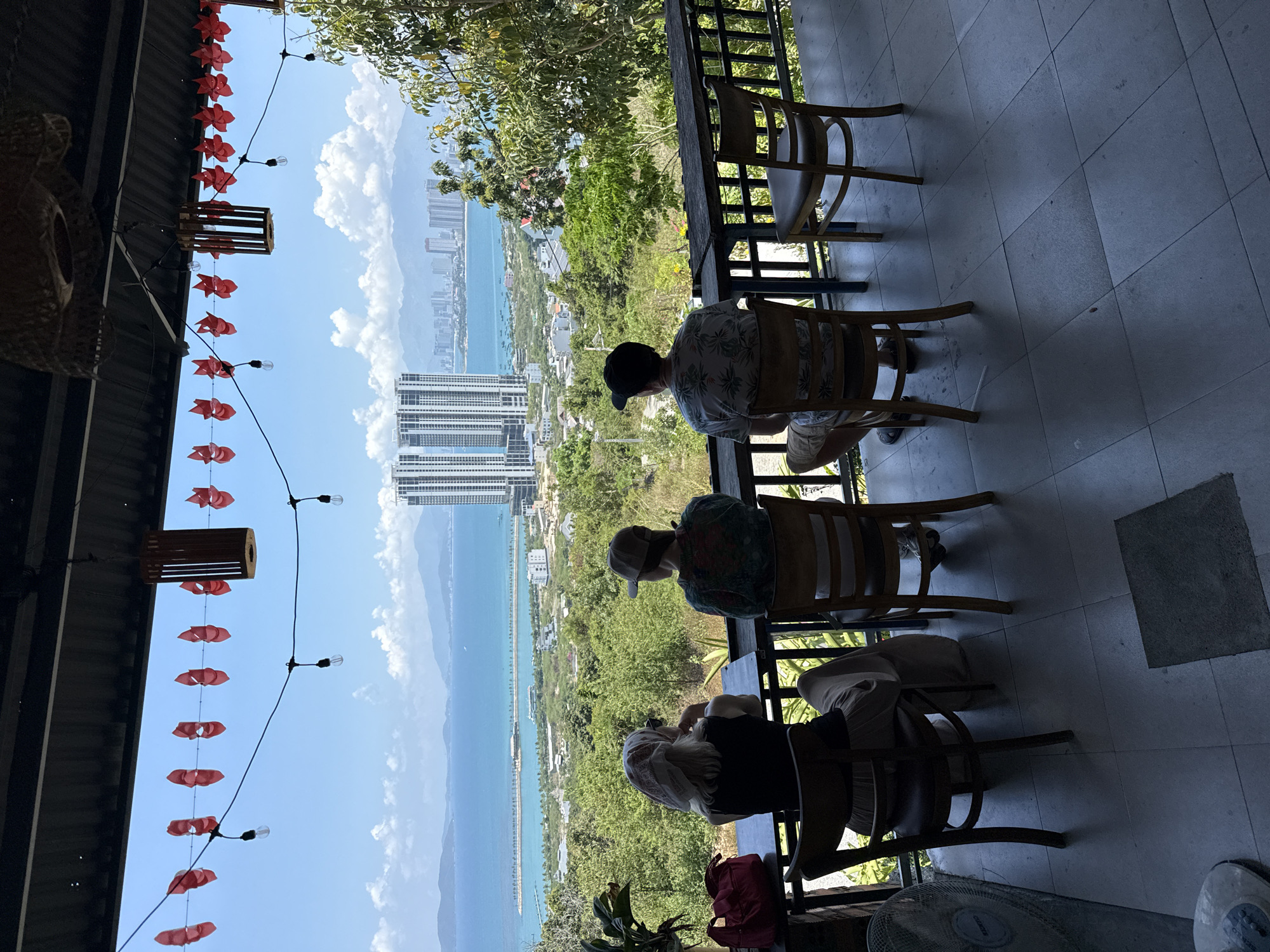 The image depicts an elevated outdoor setting, likely a cafe or restaurant balcony, offering a panoramic view of a coastal city. Three individuals are seated on wooden chairs with their backs to the viewer, facing the expansive vista. They appear to be relaxing and observing the scenery.
The foreground features a tiled floor and a sturdy wooden railing or table along the edge. Overhead, a string of decorative red elements, resembling stylized fish or flowers, is strung across the space, accompanied by small light bulbs.
Beyond the railing, a lush green hillside slopes down towards a sprawling cityscape. Numerous modern high-rise buildings line the coast, some quite tall and densely packed. Further out, the calm blue ocean stretches to the horizon, meeting a distant landmass or mountains under a clear sky dotted with white clouds. A long, thin structure, possibly a pier or causeway, is visible extending into the water.
The overall impression is one of leisure and enjoyment of a beautiful, sunny day. The time appears to be mid-day or afternoon due to the bright sunlight and shadows. There is no discernible text visible in the image. Given the location context provided, this scene is likely in Bac Nha Trang Ward, Vietnam.
