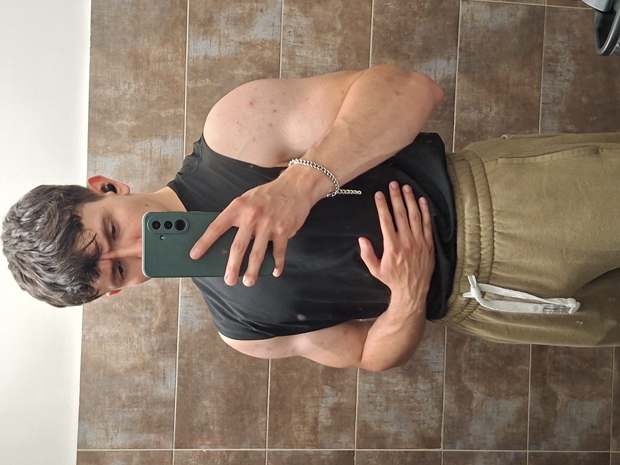 The image depicts a young adult male taking a mirror selfie in an indoor setting. The male has short, wavy, dark brown hair and is light-skinned. He is wearing a black sleeveless t-shirt and olive green or khaki sweatpants with a white drawstring visible. A silver-colored cuban link chain bracelet is on his left wrist, and a black earbud is in his left ear. Small red blemishes are visible on his right upper arm. He holds a dark green smartphone with three visible camera lenses in his left hand, partially obscuring his face. His right hand rests on his abdomen. The walls of the room are covered with rectangular, light brown or tan tiles exhibiting a distressed, somewhat mottled pattern, arranged horizontally. Above the tiled section, the upper wall or ceiling is white. No specific visual cues in the image indicate its location within the city of Plovdiv, Bulgaria. A dark object, possibly a waste bin, is partially visible in the bottom right corner.