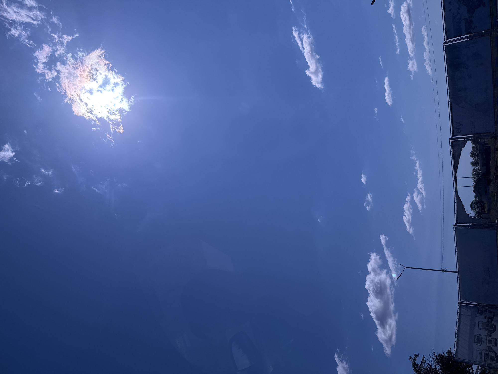 The image captures a bright, sunny sky with scattered white clouds over the city of Прилиманське, Ukraine. The sun is partially obscured by a dense cloud in the upper right quadrant, creating a radiant halo effect and lens flare. In the lower portion of the image, a segmented fence or barrier is visible, with some sections appearing damaged or torn. A tall street lamp stands near the center of the fence, its light fixture partially obscured by a small cloud. To the left of the street lamp, a dark-colored tree is partially visible. The overall scene suggests an outdoor environment with a clear sky and some urban infrastructure.