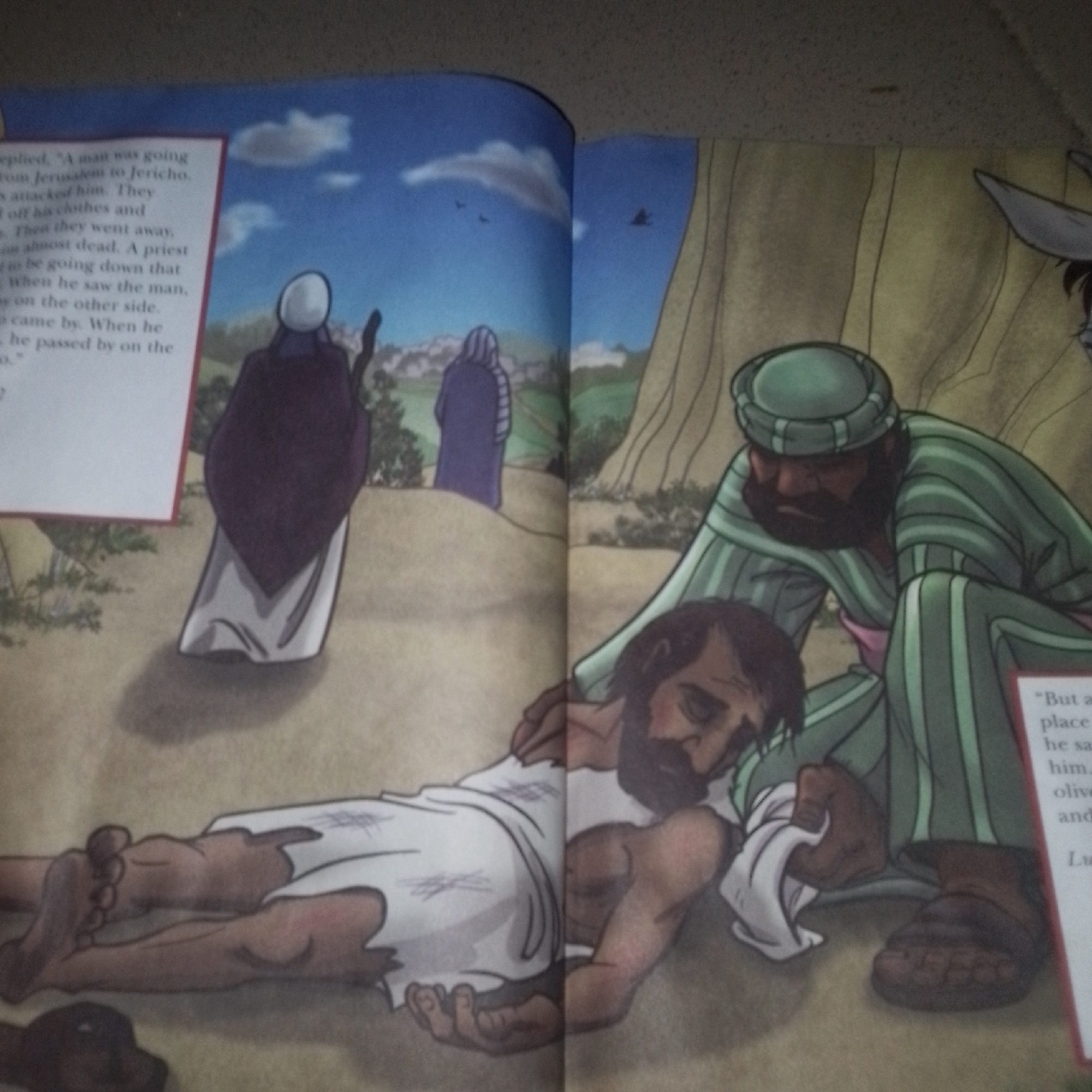 The image depicts a scene from a storybook, illustrating the Parable of the Good Samaritan. On the left, text describes a man being attacked and left for dead. Visually, the left page shows a distressed man lying on the ground, partially covered by a white cloth. His body is thin, and his feet are bare. In the background, two figures in robes are walking away, one in purple and the other in blue. The environment appears to be a landscape with rolling hills and trees.
The right page shows a man in a green and white striped robe and a green turban kneeling beside the injured man. He is tending to him, holding a white cloth. His expression is compassionate. In the far right background, the ear and head of a donkey are visible, suggesting the Samaritan arrived on a donkey. A snippet of text on the right reads, "But a Samaritan, as he journeyed, came to where he was; and when he saw him, he had compassion..." This refers to a passage from Luke 10:33.
The image does not contain any information about the city of Jalingo, Nigeria.