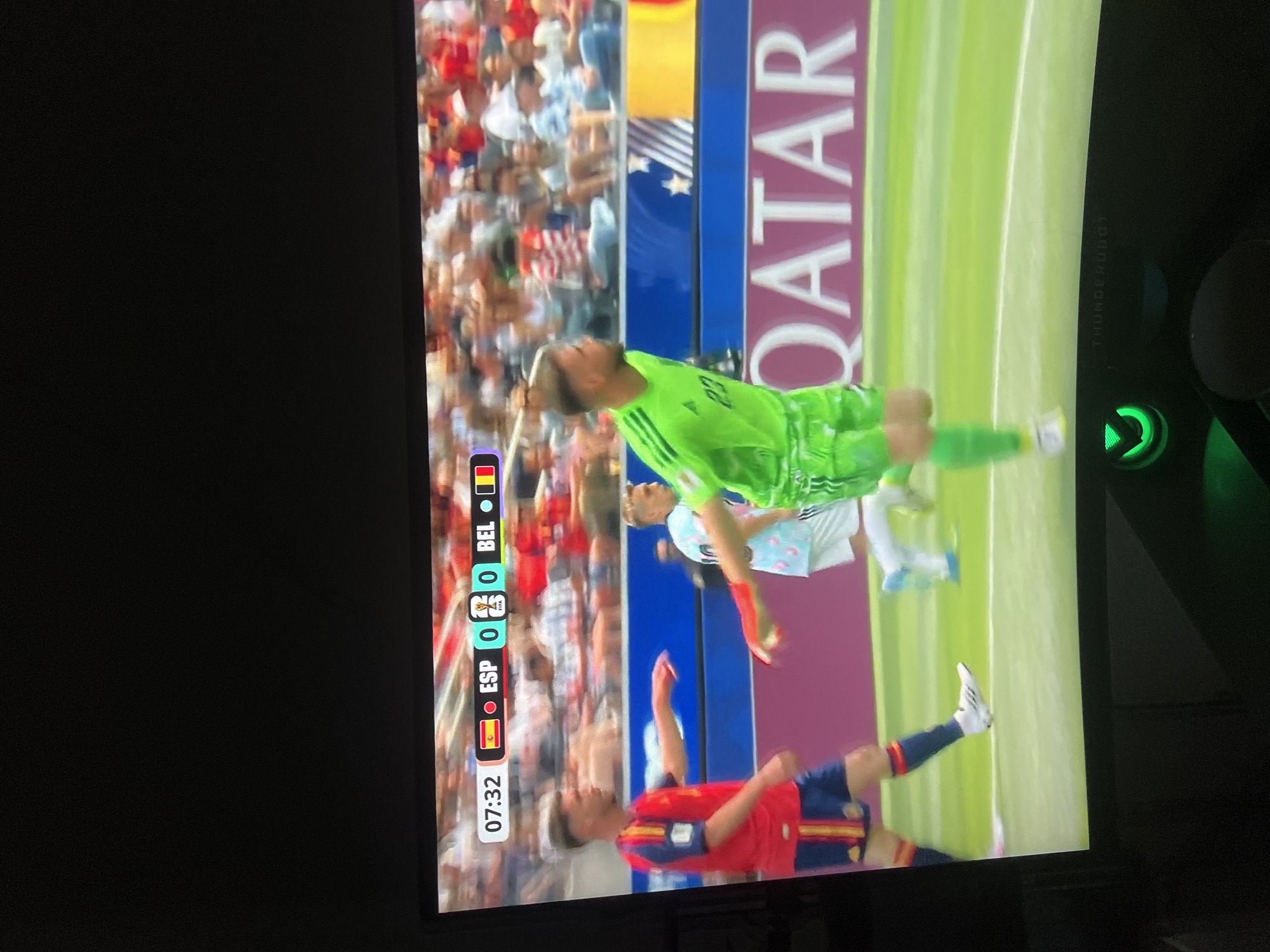 A screen displays a soccer match with the score of Spain 0 and Belgium 0 at 7 minutes and 32 seconds into the game. A player in a red and blue Spanish uniform with the number 10 on his sleeve is positioned on the left side of the frame, facing right with his left arm outstretched. To his right, a goalkeeper in a bright green uniform with the number 23 on his chest is running and looking left. Spectators are visible in the blurred background behind a blue barrier with stars and a banner that reads "QATAR". The screen is tilted slightly upwards, and a green illuminated logo and the text "THUNDEROBOT" are visible on the bottom bezel of the screen. The image is taken indoors and captures a portion of a dark room at the bottom, suggesting the screen is part of a display device. The location of Sinyavino, Russia is not depicted in this image.