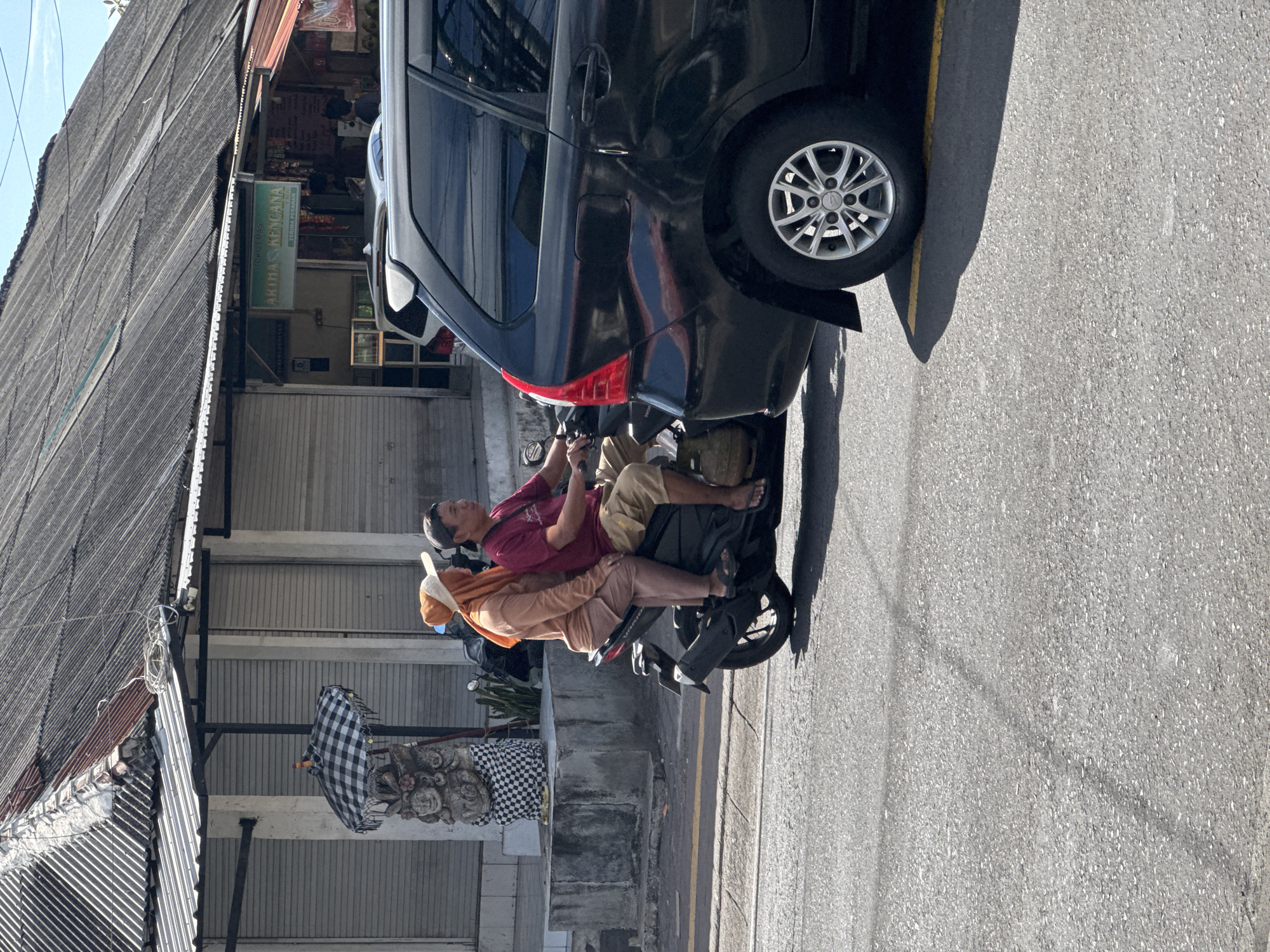 The image shows a street scene in Kuta Selatan, Indonesia. A black car is parked on the side of the road, partially blocking a motorbike. On the motorbike, two people are visible: a man in a red shirt and beige shorts, and a woman in an orange headscarf and long-sleeved shirt. Both appear to be looking in the same direction. The background consists of buildings with corrugated metal roofs and storefronts. A checkered umbrella stands next to a stone sculpture on the sidewalk. The road is paved and appears to be in good condition.