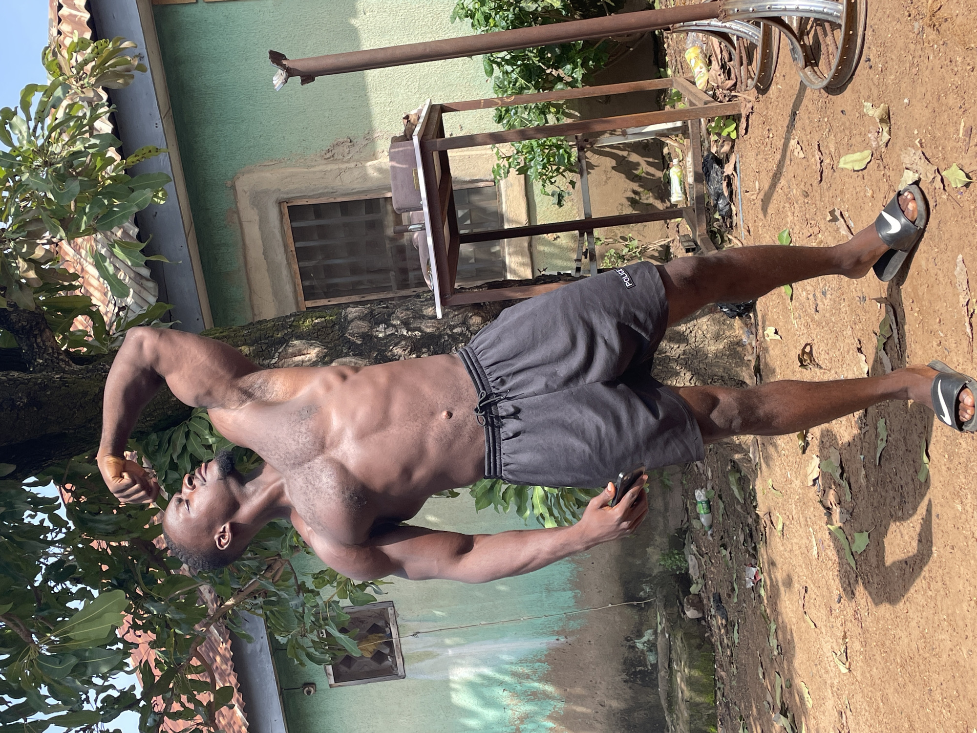A shirtless Black male, appearing to be in his 20s or 30s, is posing outdoors. He has short black hair, a goatee, and a muscular physique. He is wearing black athletic shorts with the word "POLICE" printed in white on the left leg, and black slide sandals with white Nike logos. He is standing next to a tree with his right arm flexed upwards, showing his bicep. Behind him is a light green building with a corrugated metal roof and a window with bars. A metal table with objects on it, and a rusty metal structure with wheels are also visible. The ground is dirt covered with fallen leaves. The overall impression is that the photograph was taken in a residential area, possibly in the city of Akwanga, Nigeria, given the architectural style of the building and the surrounding vegetation.