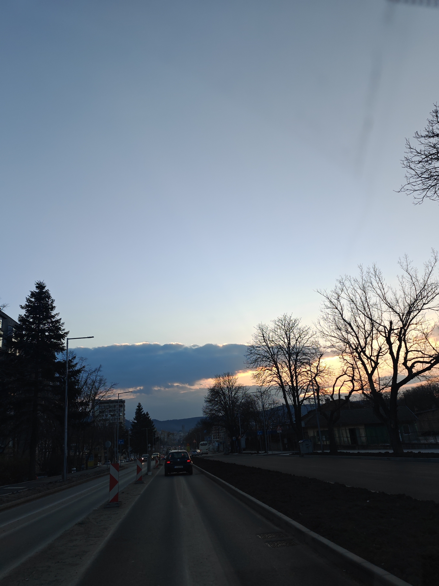 The image depicts an urban street scene in Shumen, Bulgaria, at dusk. The sky transitions from a light blue overhead to an orange-yellow hue at the western horizon, partially obscured by a band of dark, low-lying clouds. A multi-lane asphalt road extends into the distance, with two lanes visible heading away from the viewer. A dark sedan with illuminated taillights, bearing Bulgarian license plate H 0048XC, is positioned in the right lane, moving away from the camera. The right shoulder of the road shows evidence of recent excavation or construction, with disturbed dark earth and two red-and-white striped chevron traffic markers delineating the work zone. The left side of the road features a paved shoulder, a streetlamp, and an evergreen fir tree. Silhouetted deciduous trees with bare branches line both sides of the street, particularly prominent against the illuminated sky on the right. Various urban buildings, including multi-story residential blocks and smaller, older structures, are visible in the background among the trees. Distant vehicle headlights are observable further down the road. Hills or mountains are faintly visible on the distant horizon.