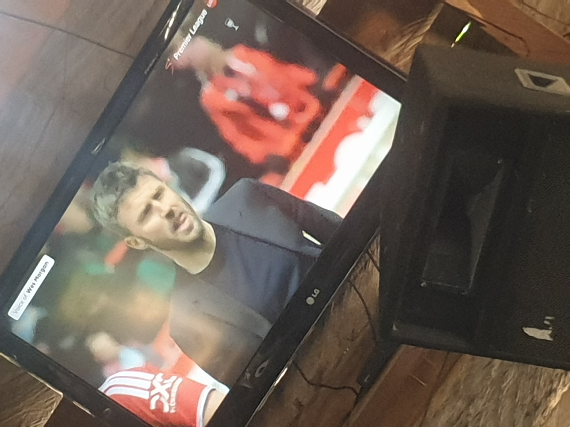 The image shows a television screen displaying a Premier League soccer broadcast. A man in a dark shirt is visible on the screen, likely a player or commentator, and text on the screen identifies the “Voice of Wes Morgan”. The TV is an LG model. The setting seems to be indoors, and the TV appears to be set on a rough, possibly unfinished wooden surface. An unidentified black object partially obscures the right side of the television. There are no indicators of a specific city or location.