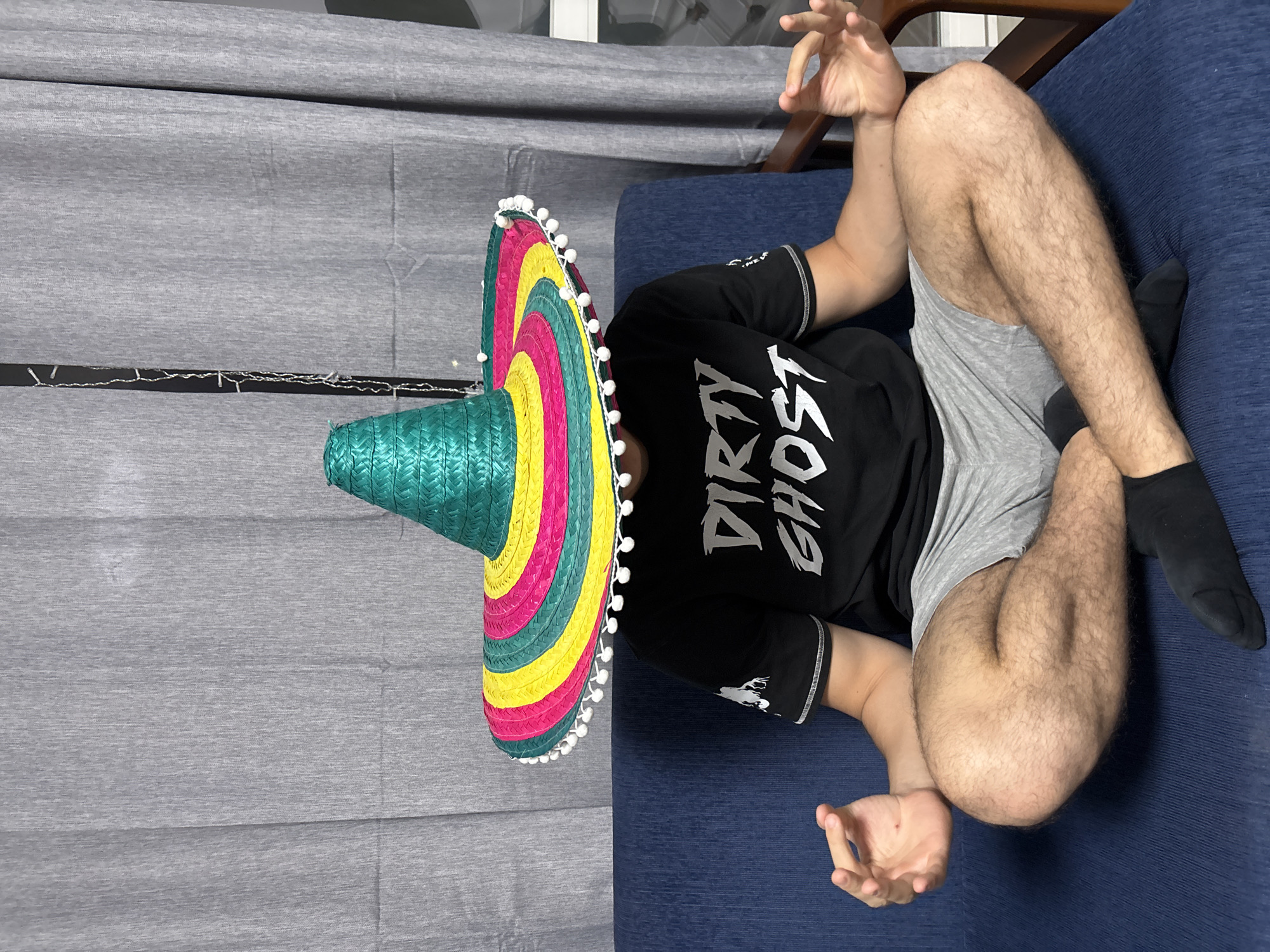 This image depicts a person lying on their back on a blue upholstered couch or chair within an indoor setting. A large, vibrant sombrero completely covers the person's head and face. The sombrero features a teal conical crown and a wide brim decorated with concentric rings of teal, pink, yellow, and green, finished with a border of small white pom-poms.
The person is wearing a black t-shirt with the white, stylized text "DIRTY GHOST" prominently displayed on the front. They are also dressed in grey shorts and black socks. Their arms and legs are visible, showing body hair, with their knees bent and hands resting in slightly open, relaxed gestures.
The background consists of grey fabric curtains or wall coverings. A string of small white fairy lights is visible across the grey surface. The scene suggests a casual, relaxed home environment, possibly a living room or bedroom. There are no specific visual cues within the image to identify the location as Plovdiv, Bulgaria.