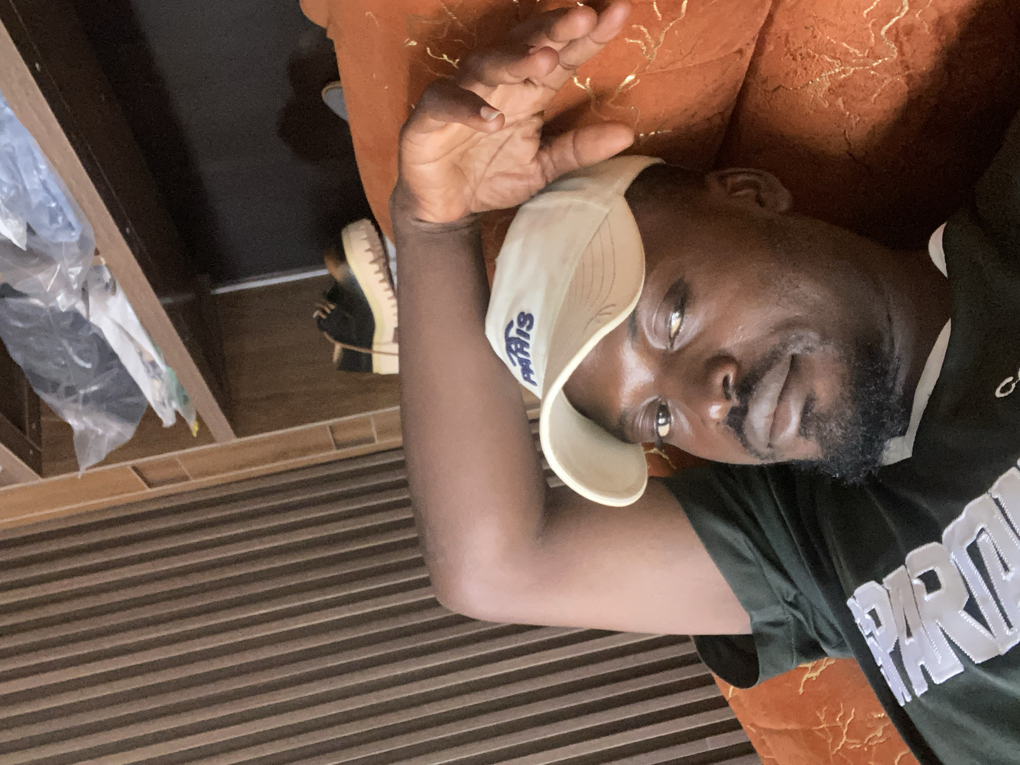 A man wearing a black t-shirt with white lettering and a white baseball cap with blue lettering is lying on an orange couch. The man has a beard and is looking towards the camera. Behind him, a wall with vertical wooden slats is visible, and above that, a shelf holds various items, including plastic bags and a pair of black and tan platform shoes. The scene appears to be indoors, likely in a residential setting in Gura Top, Nigeria.