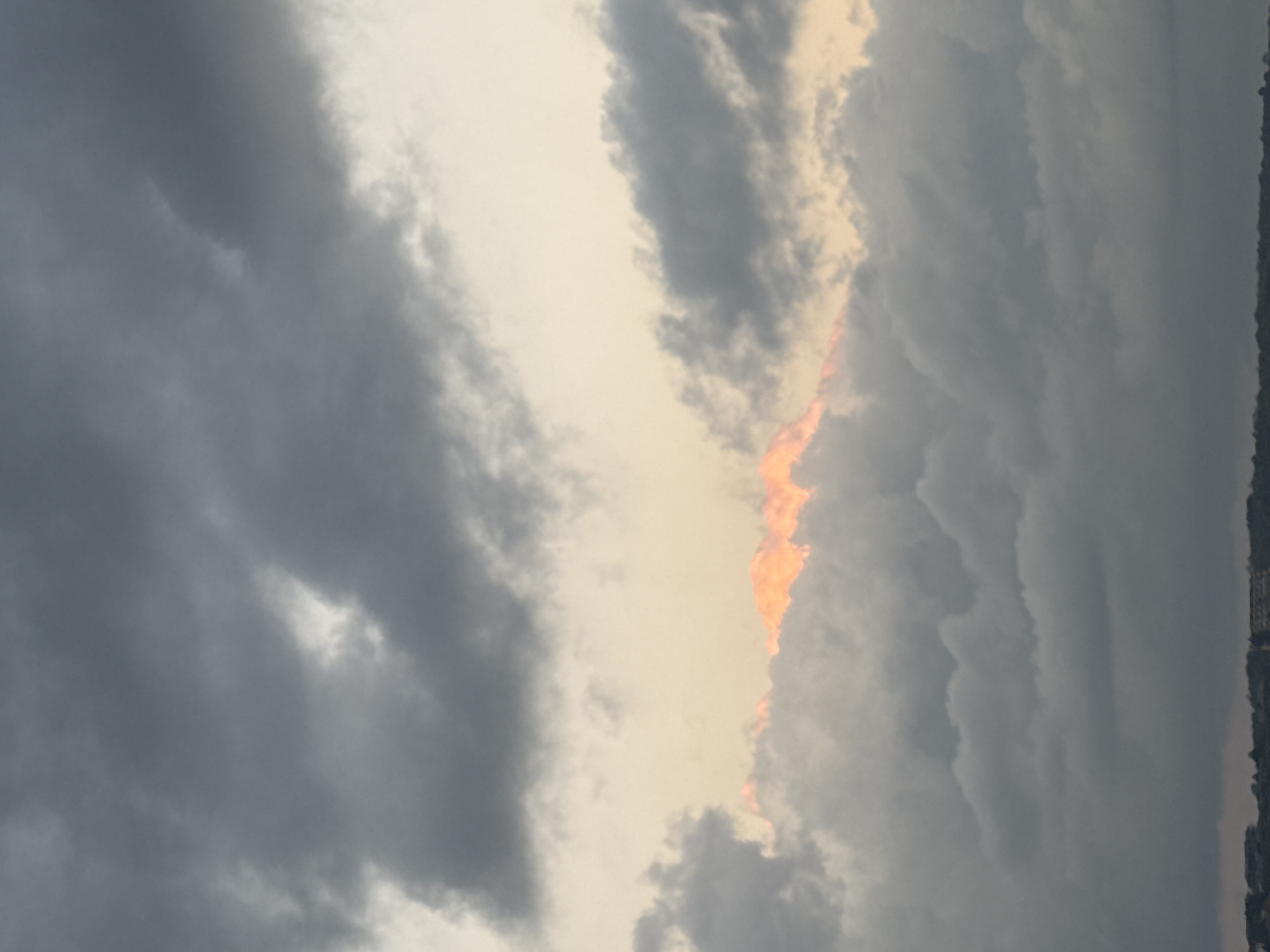 The image depicts a cloudy sky with a sliver of orange-pink light peeking through the clouds in the middle section. The lower portion of the image shows a dark silhouette of a treeline and some buildings, suggesting an urban or coastal landscape. This scene is located in Kuta, Indonesia.