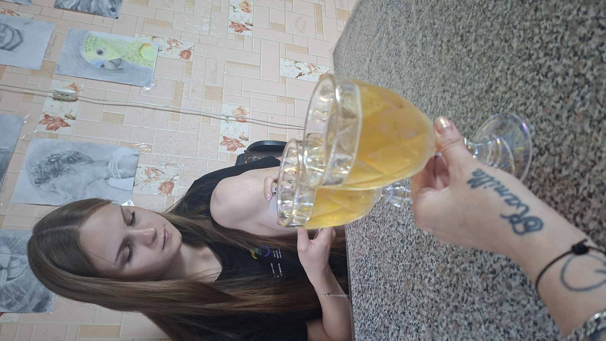 A young woman with long brown hair is sitting at a table, looking down at two glasses of amber liquid. She is wearing a black t-shirt. Her left arm is resting on her knee, and her right hand is reaching out to hold one of the glasses. On her left forearm, a tattoo of the name "Kamilla" is visible. The table is covered with a speckled, granite-like material. In the background, drawings of a lion, a person's profile, and a parrot are taped to a wall with a patterned wallpaper. The scene appears to be indoors, likely in a residential setting in Myadzyel, Belarus.