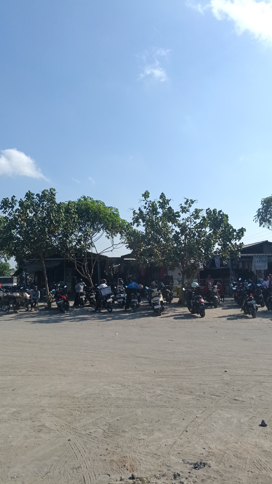 The image depicts a bright, sunny day in Dukuh, Indonesia, with a clear blue sky. The foreground shows a dusty, unpaved parking area with tire tracks visible in the dirt. In the middle ground, a row of motorcycles is parked, some with riders seated on them, suggesting a temporary stop. Behind the motorcycles, a line of small buildings or stalls is visible, with trees providing shade. A sign with the word "LERVIA" is visible on one of the buildings. The scene appears to be a typical outdoor marketplace or rest stop, possibly near a road or tourist attraction.