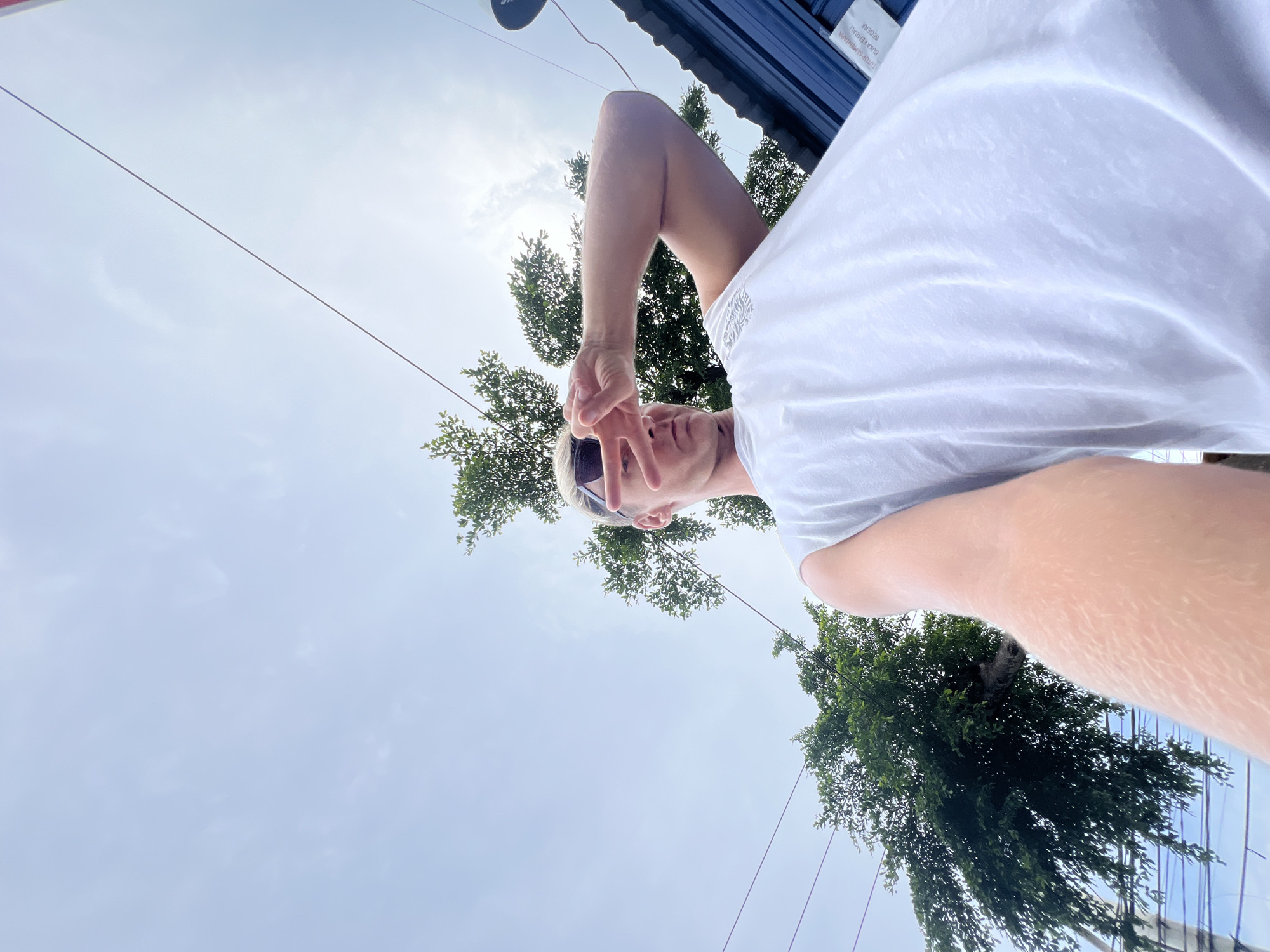 An outdoor, upward-angle selfie depicts a fair-skinned male subject with light-colored hair and dark sunglasses, wearing a white sleeveless shirt. He holds the camera with his right arm extended. His left hand is raised to his face, with two fingers forming a "V" shape, partially obscuring his face and sunglasses, seemingly shielding his eyes from bright overhead light. The sky is light blue with scattered clouds, and the sun appears as a glare behind the subject's head. Green trees are visible in the background against the sky. Multiple black utility lines traverse the upper and mid-frame. To the right, a dark blue corrugated metal structure is visible with a white sign attached, displaying partially legible Indonesian text: "TUTUP SEMENTARA" (Temporarily Closed) and "BUKA KEMBALI SEGERA" (Reopen Soon). This scene is located in the city of Celuk, Indonesia.