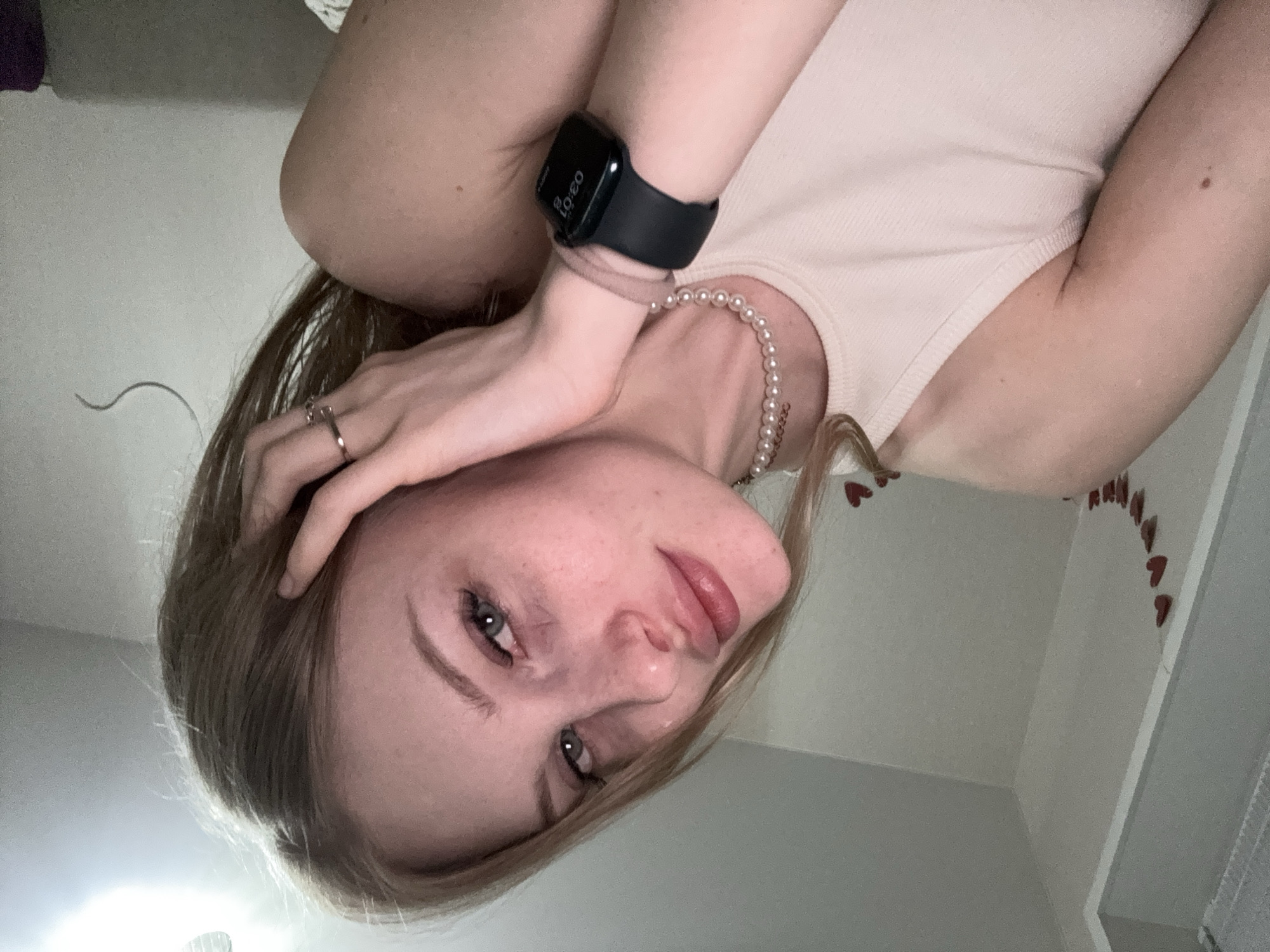 A young woman with blonde hair and blue eyes poses for a selfie in a room. She is wearing a light pink tank top and a pearl necklace. On her wrist, she wears a black smartwatch. The background consists of a plain wall with a string of red hearts pinned to it. The lighting appears to be artificial, casting a soft glow on the scene. There is no identifiable information about the location within the city of Perm, Russia.