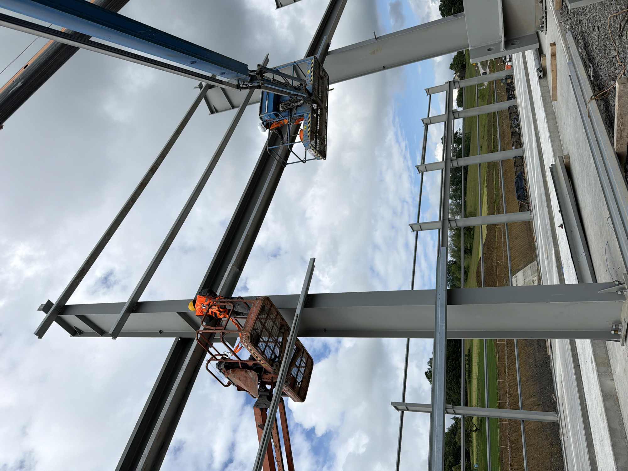 The image depicts a construction site in Stróża, Poland, featuring the erection of a steel-framed building under a cloudy sky. Two construction workers, each wearing a high-visibility orange vest and a hard hat, are operating from elevated platforms. The worker on the left is in an orange boom lift, positioned to attach a horizontal steel beam to a vertical I-beam. The worker on the right is in a blue boom lift, also engaged in work on the steel structure. The surrounding environment shows a sloped, excavated area with exposed earth and some vegetation in the background, suggesting ongoing earthworks. Concrete slabs are laid out in the foreground, with additional steel components scattered on the ground, indicating the early stages of construction.